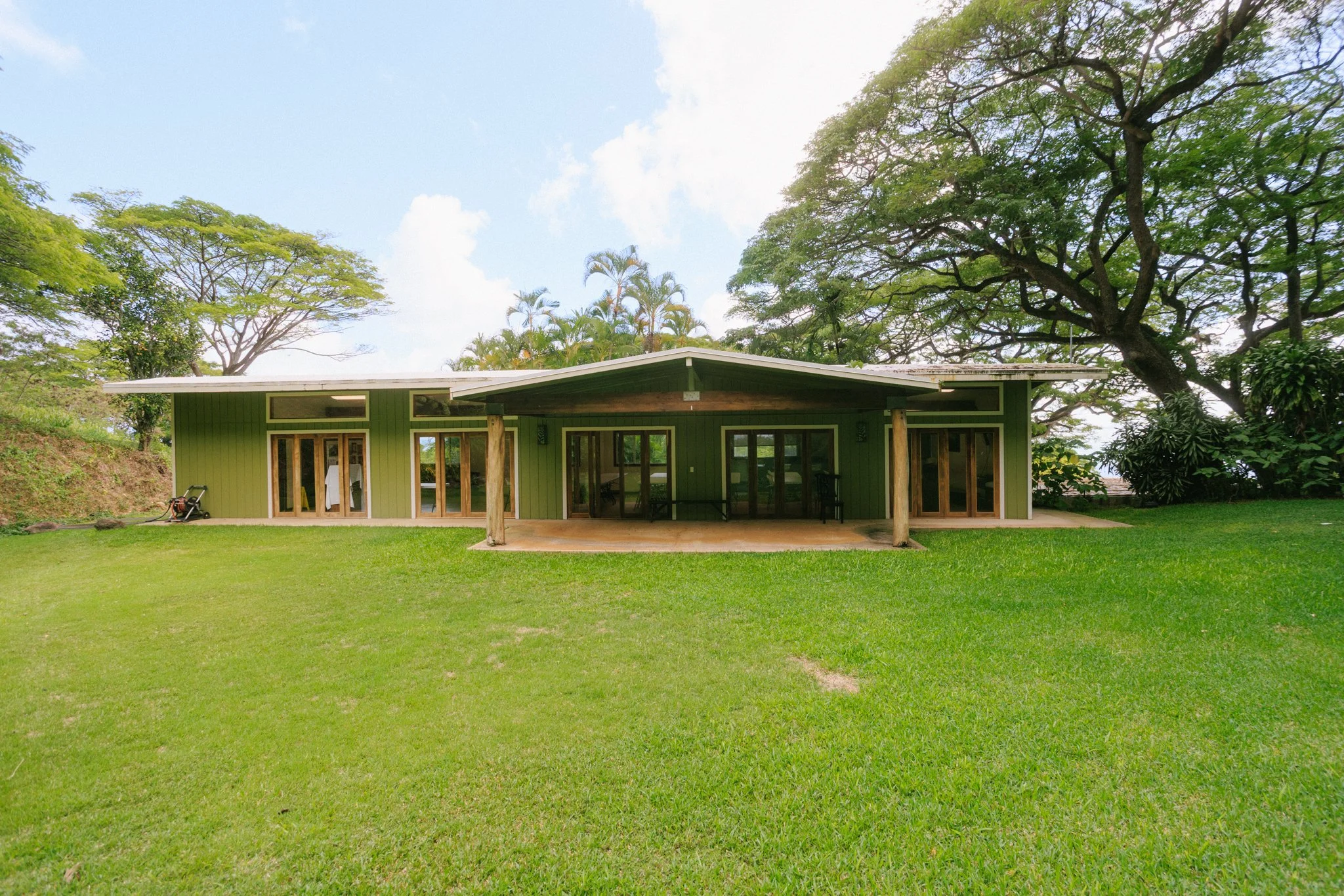 A single-story green house with large windows and sliding glass doors, surrounded by green lawn and tall trees, under a partly cloudy sky.