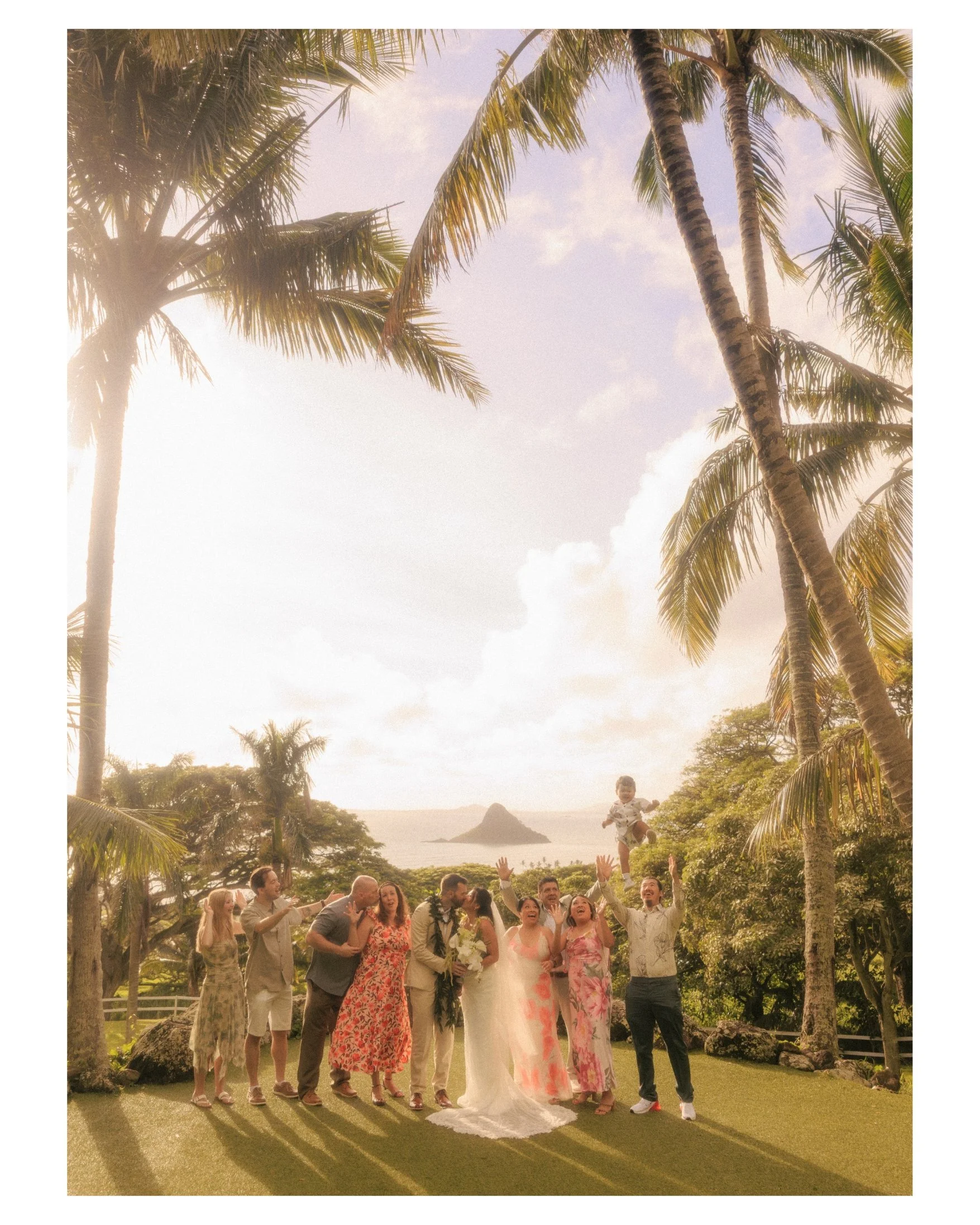 Nicole and Ethan pose with all of their wedding guests in a group portrait under leaning palm trees at Paliku Gardens, Kualoa Ranch, with sunlight flaring through the trees and Mokoli'i island in the distance on Oahu, Hawaii.