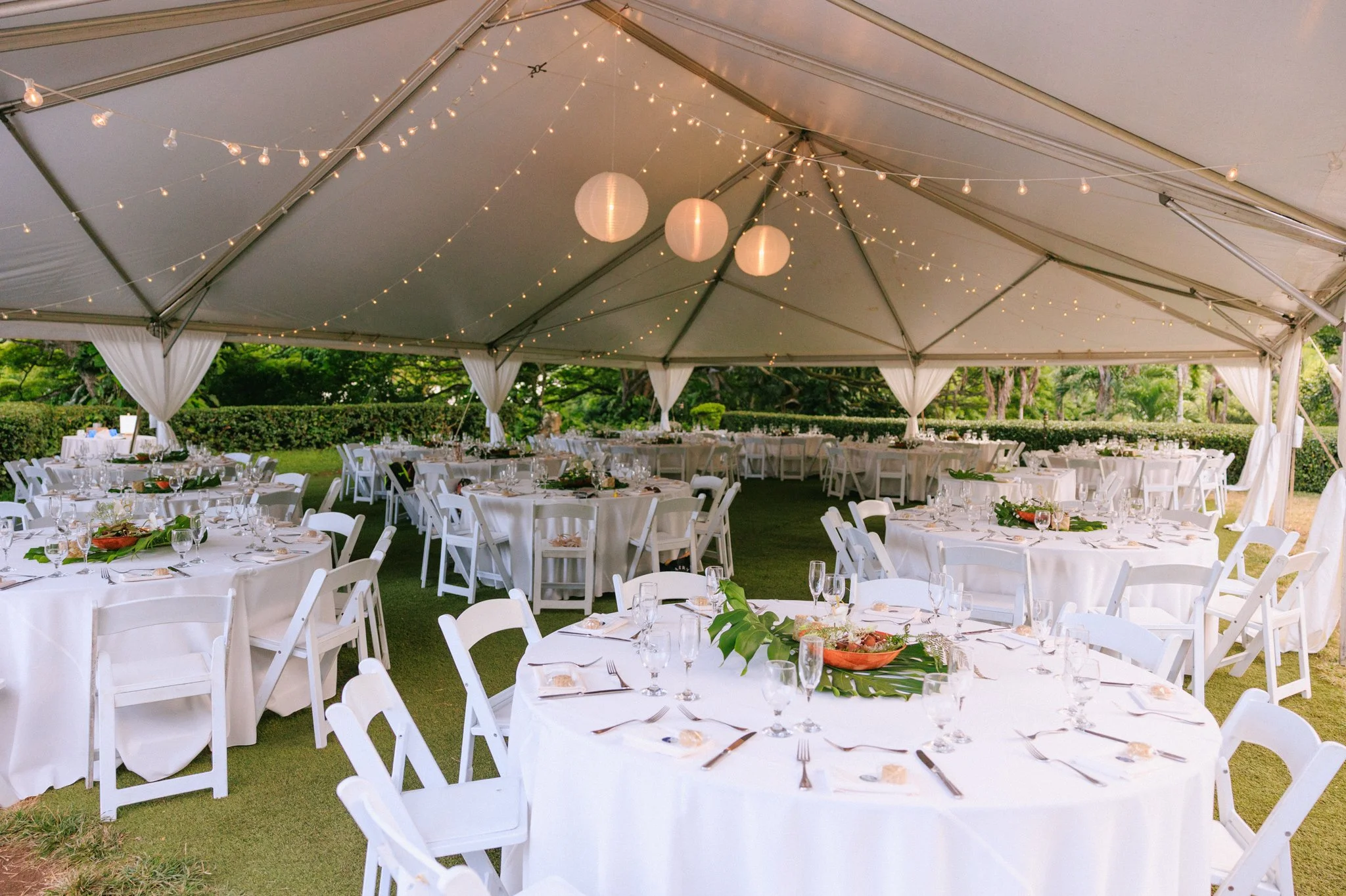 Decorated outdoor event tent with white tables and chairs, string lights, and hanging paper lanterns, set up for a celebration or wedding reception.