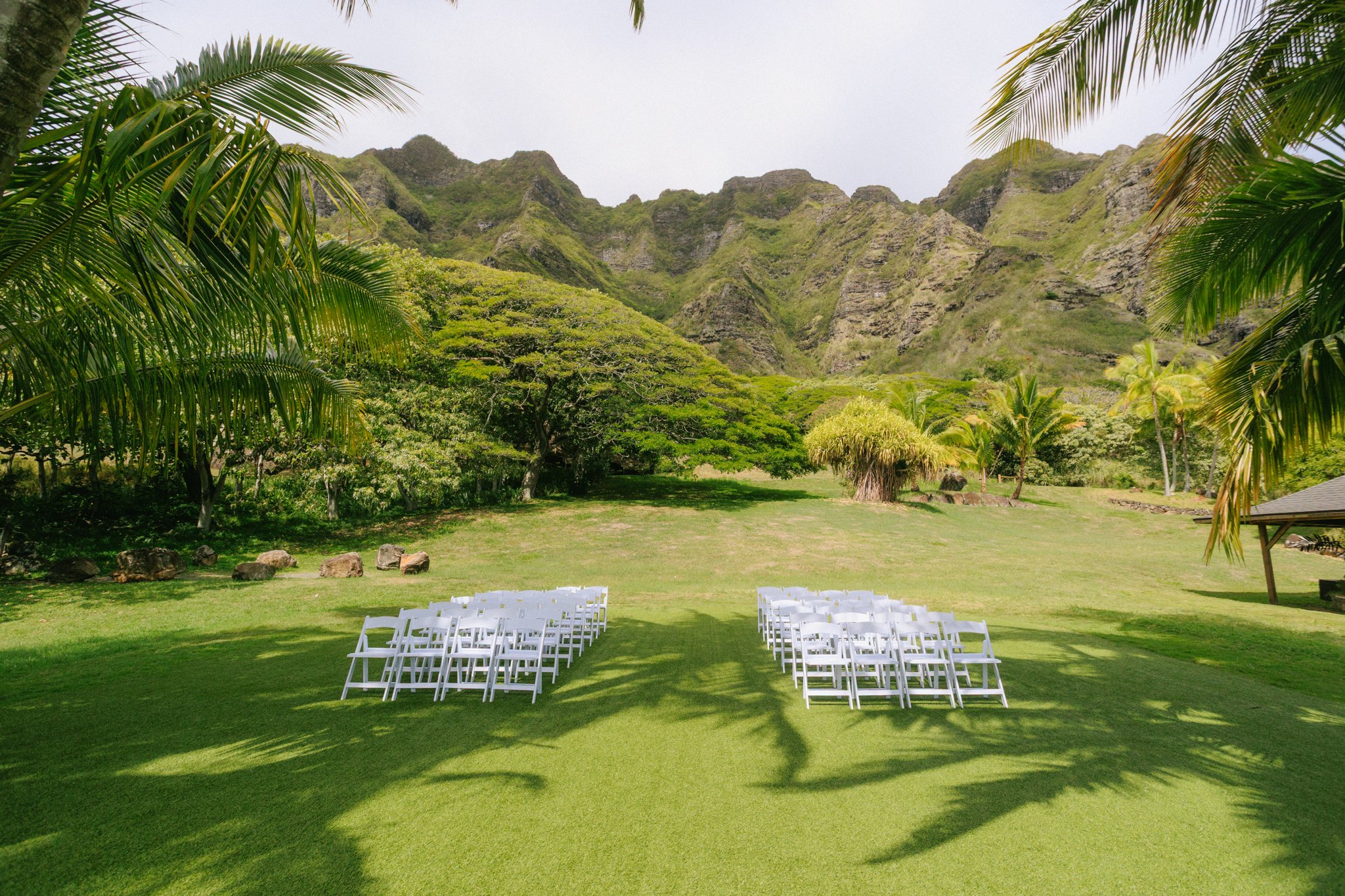 Outdoor wedding ceremony setup with white chairs on a grassy field, surrounded by tropical trees and mountains in the background.
