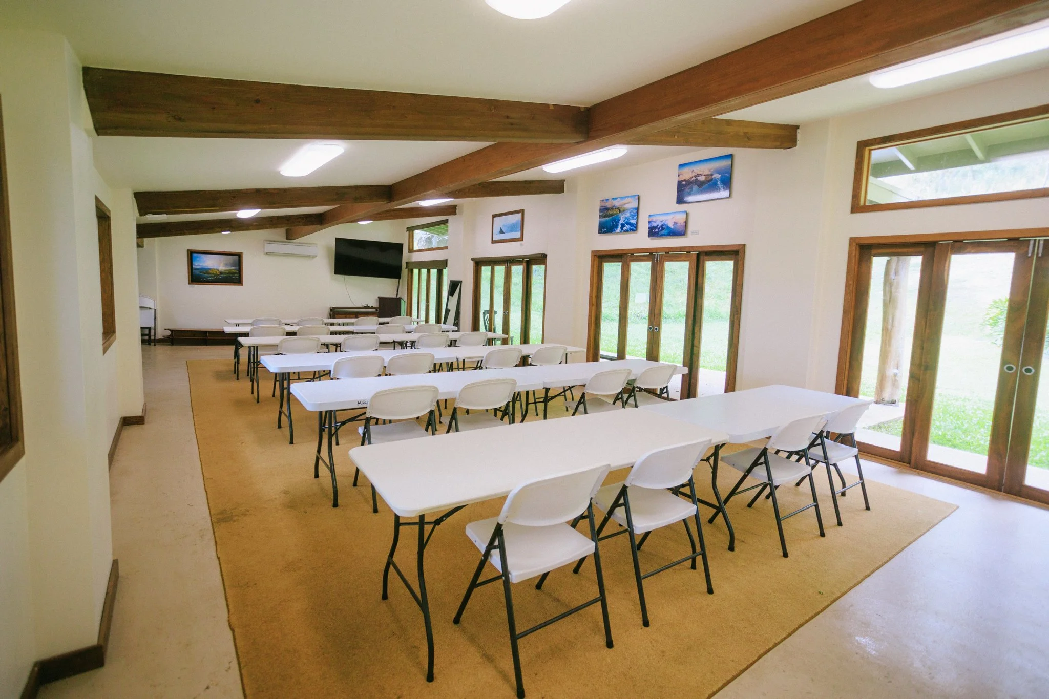 A spacious conference room with white tables and matching white chairs, large windows with wooden frames, and landscape photographs on the walls.