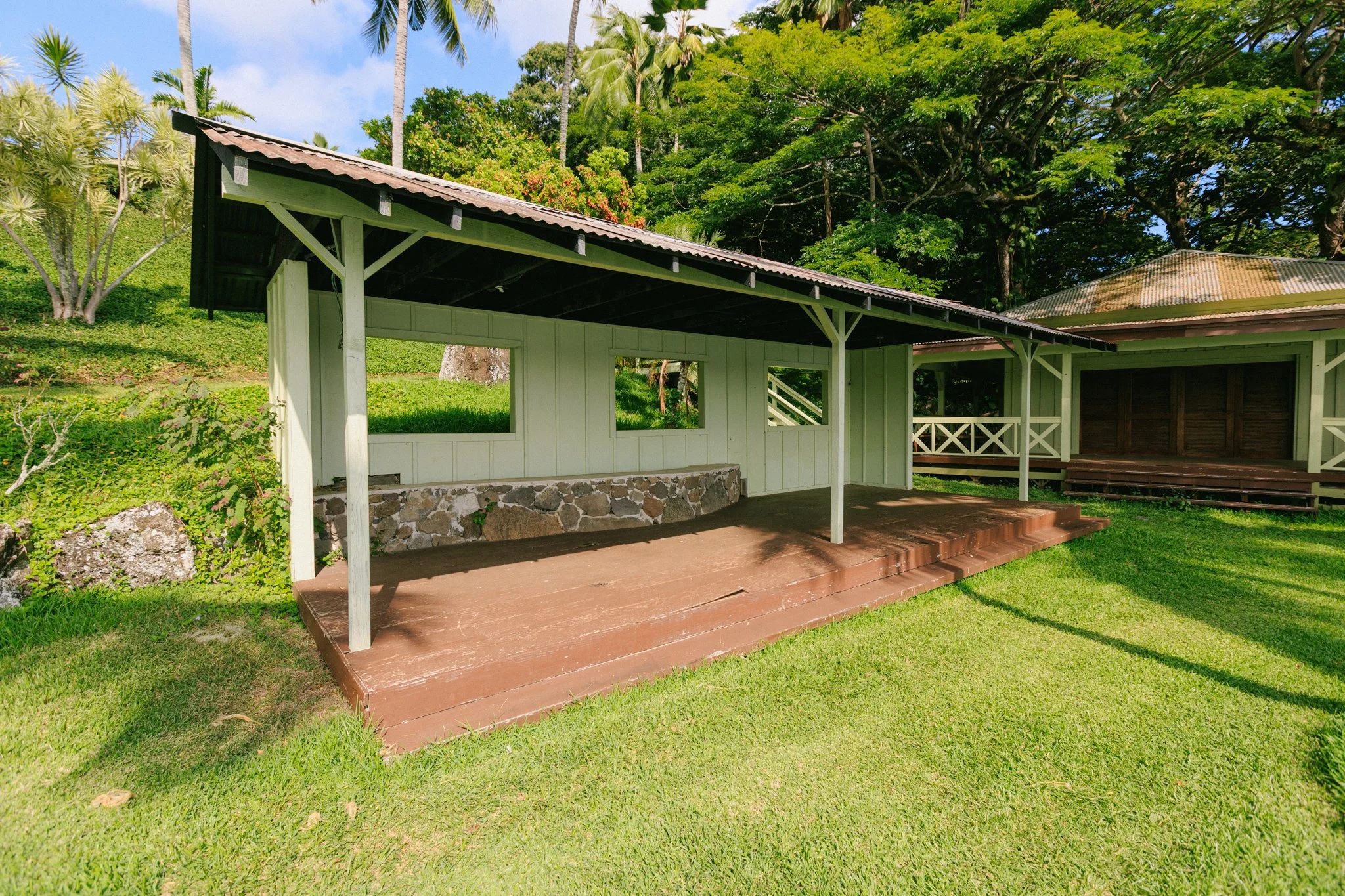 A small, open wooden pavilion with a stone foundation and corrugated metal roof, situated on a grassy area with trees and a hill in the background.