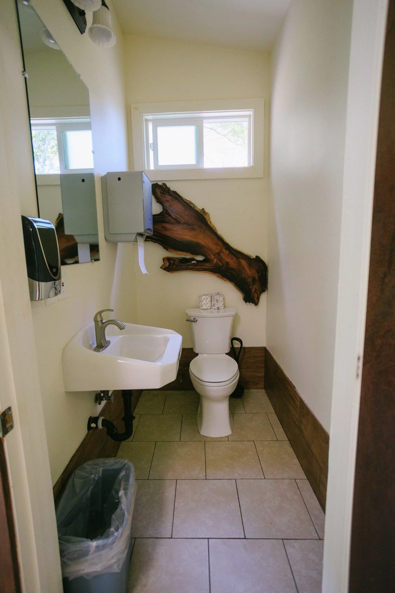 A small bathroom with a toilet, a wall-mounted sink, a mirror, a soap dispenser, a window, and a large piece of wood art on the wall.