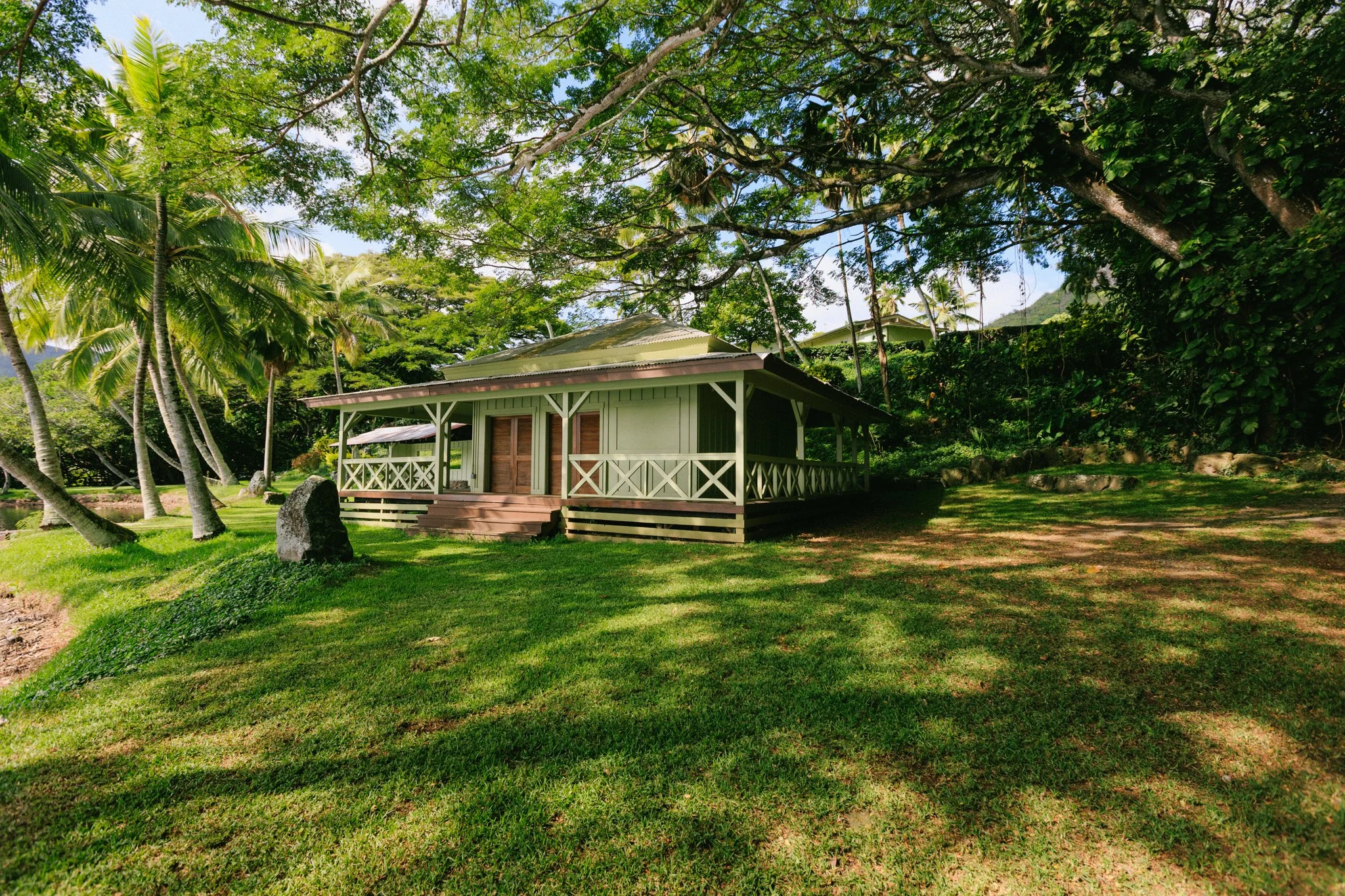 A small house in a lush, green outdoor setting with tall trees, vibrant grass, and a stone on the ground in front.