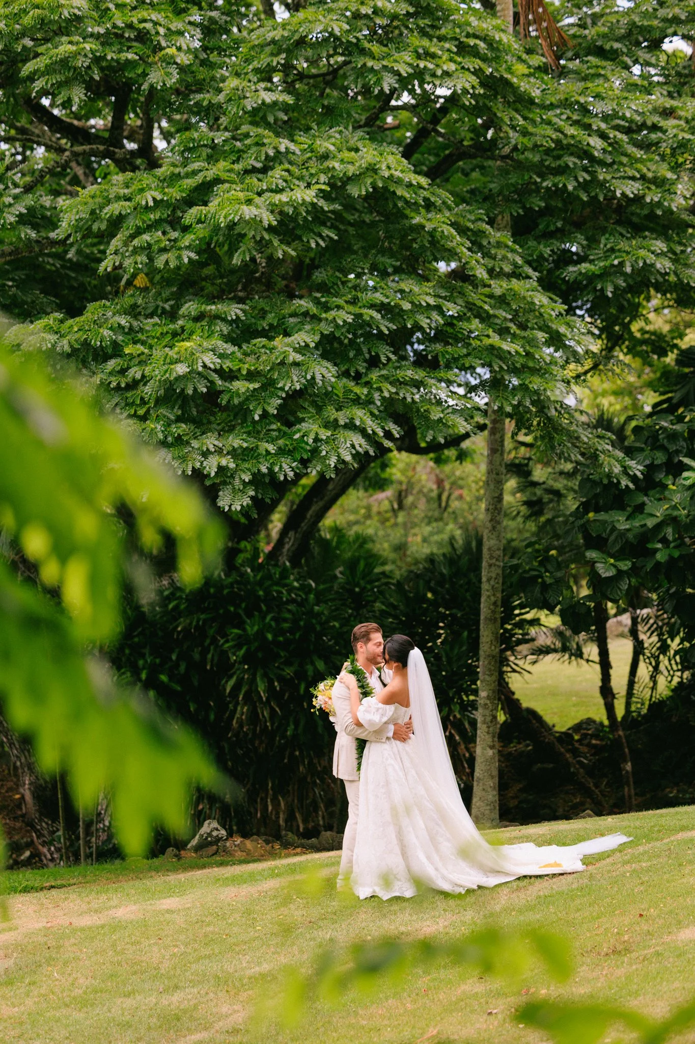 A bride and groom standing outdoors, embracing and looking at each other, surrounded by lush green trees and grass.