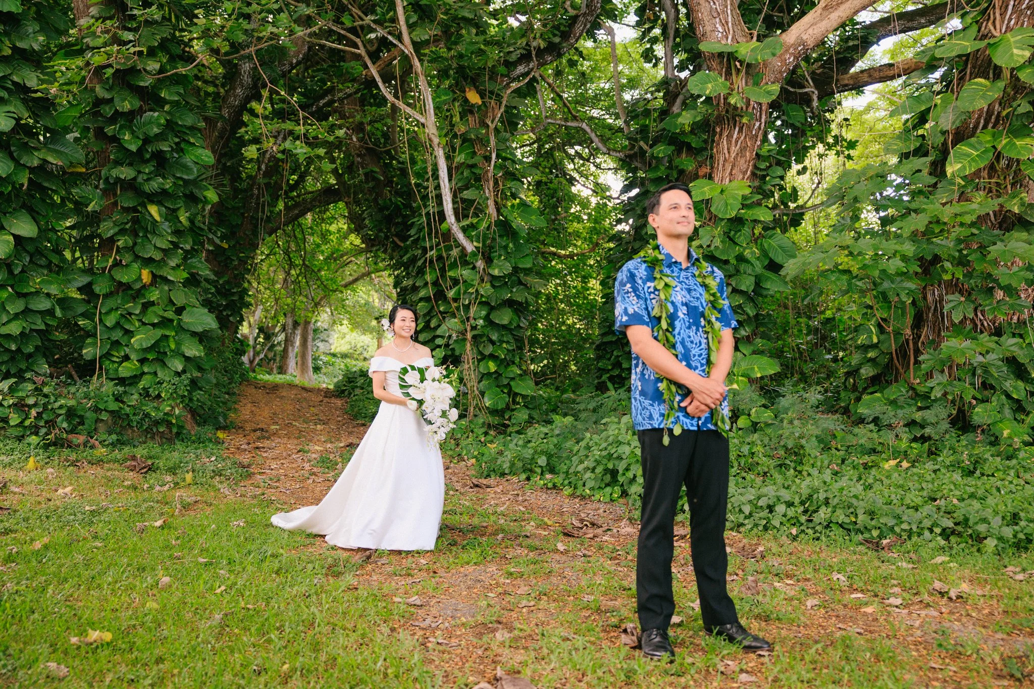 A couple stands outdoors in a lush forest, with the woman in a white wedding dress holding a bouquet, and the man in a blue floral shirt and black pants, wearing a green lei around his neck.