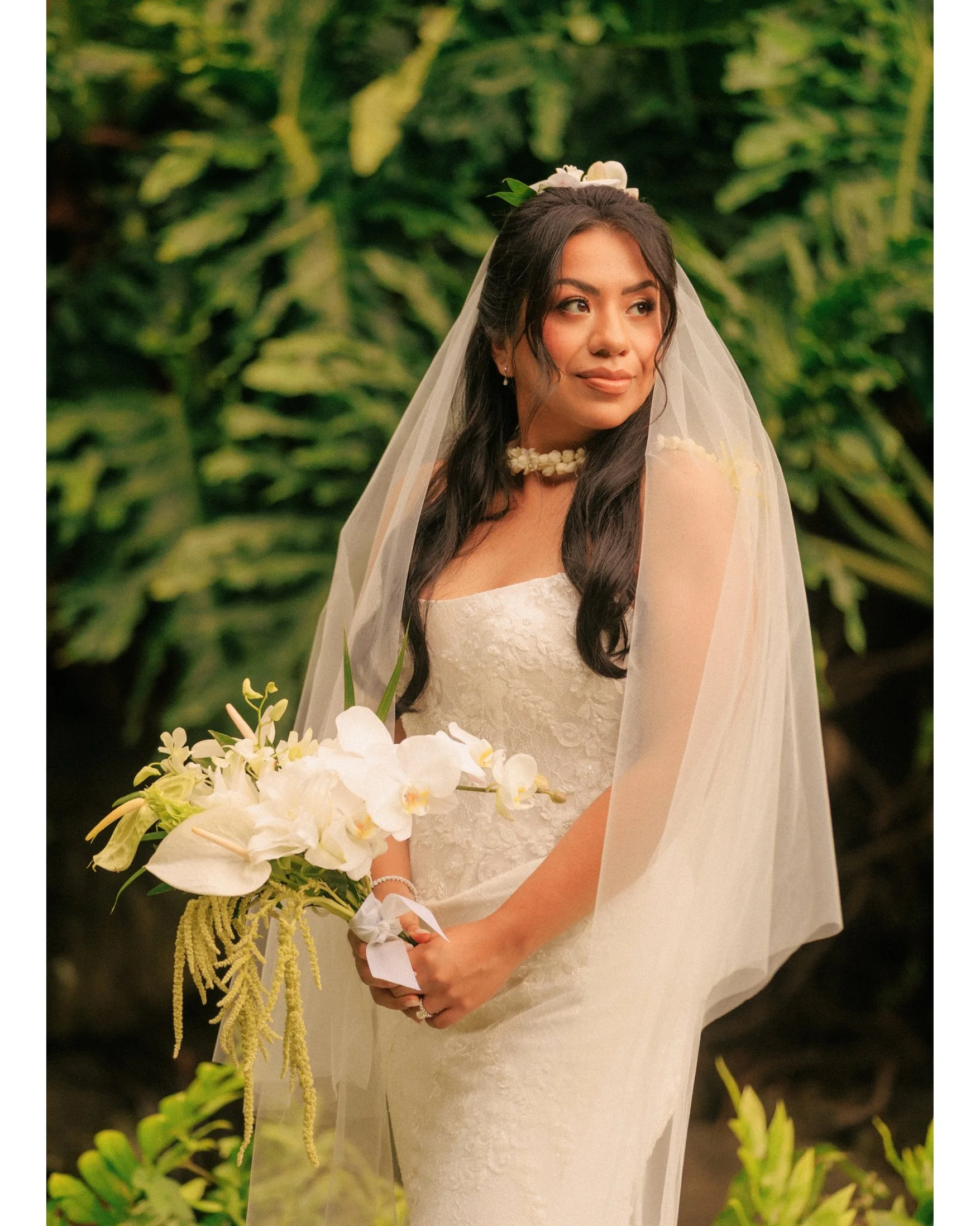 Nicole stands alone in her white dress and veil holding a white orchid bouquet against lush green foliage at Kualoa Ranch on Oahu, Hawaii, in a soft bridal portrait from her morning elopement.