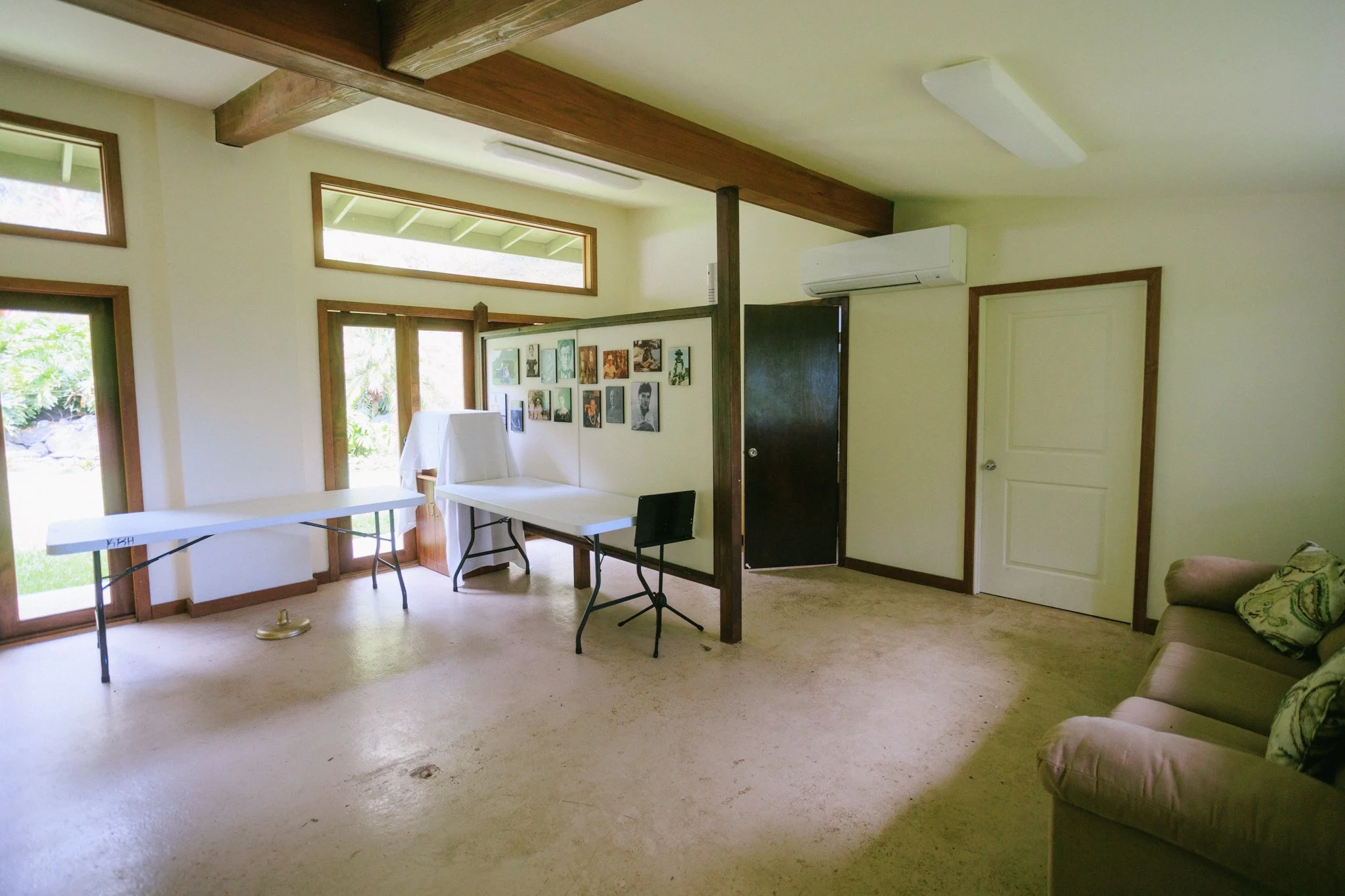 An interior room with windows and a door, with a beige carpeted floor, a beige sofa with patterned pillows, a white table, and a wall with a gallery of framed pictures. There are ceiling lights, an air conditioning unit, and a partial view of a patio