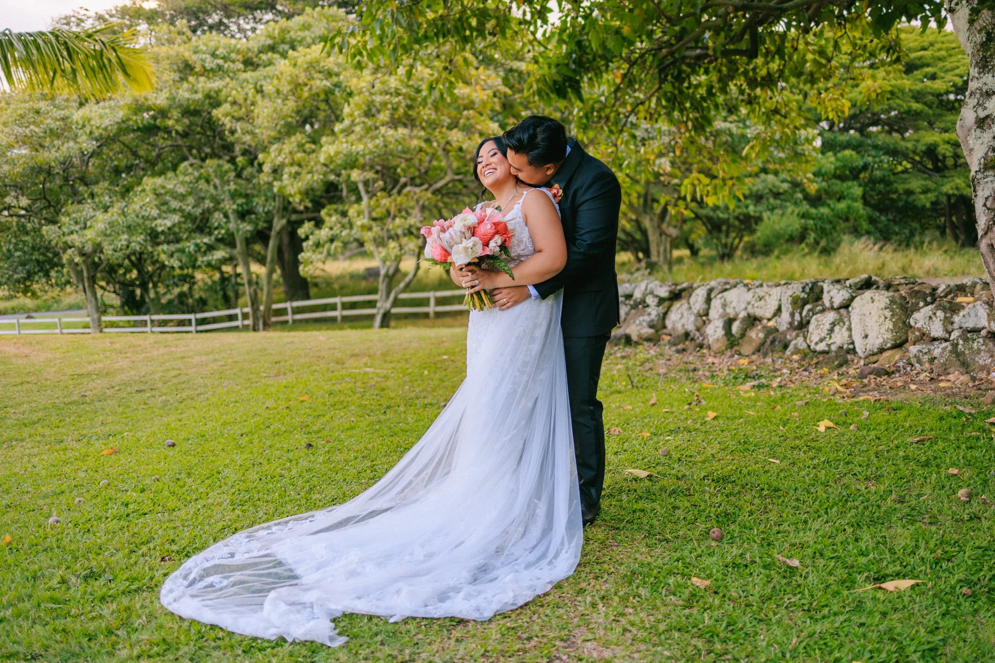 A bride in a white wedding gown holding a bouquet of pink and white roses, and a groom in a black suit, sharing a kiss outdoors with green trees and a stone wall in the background.