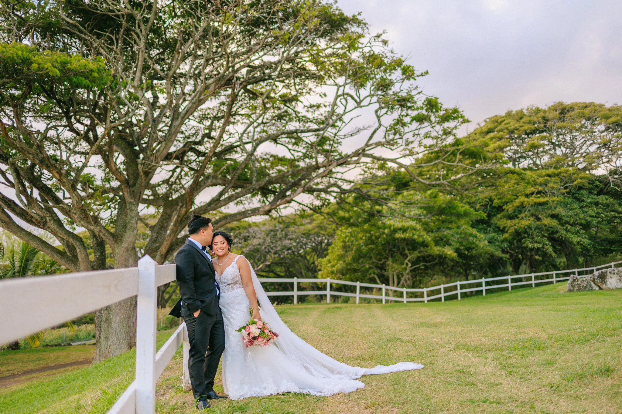 A bride and groom standing close together outdoors on a grassy area, with trees and a white fence in the background. The bride is holding a bouquet of pink and white flowers, and both are dressed in formal wedding attire. The bride is smiling with he