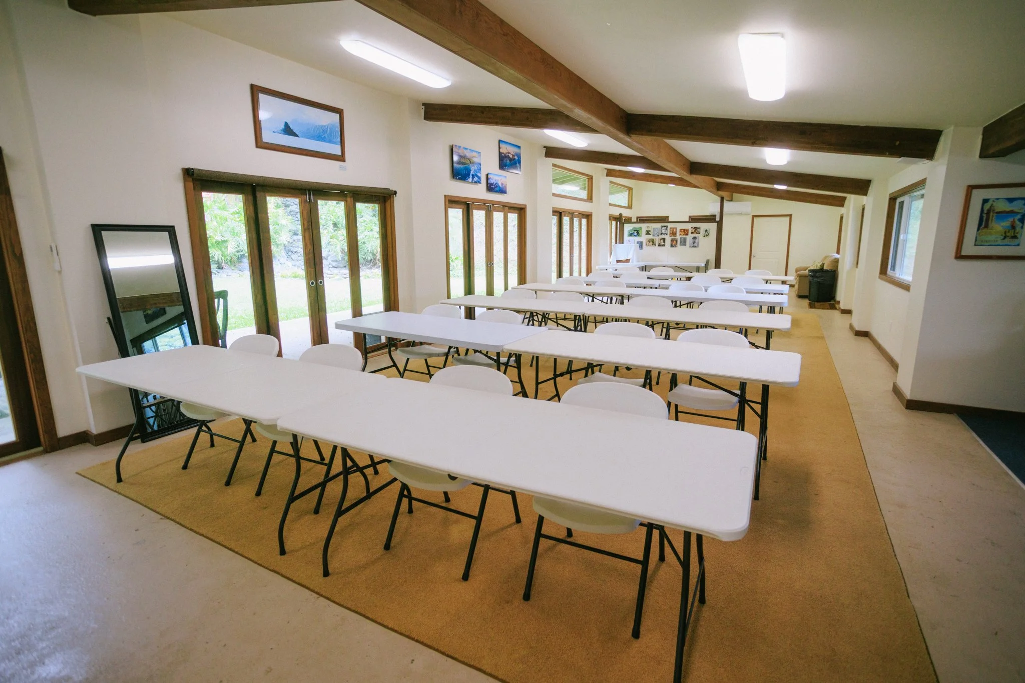 A bright, spacious room with white walls and a sloped ceiling featuring wooden beams. There are several long white tables arranged in rows, surrounded by white chairs with black legs. Large glass sliding doors on one side let in natural light and rev