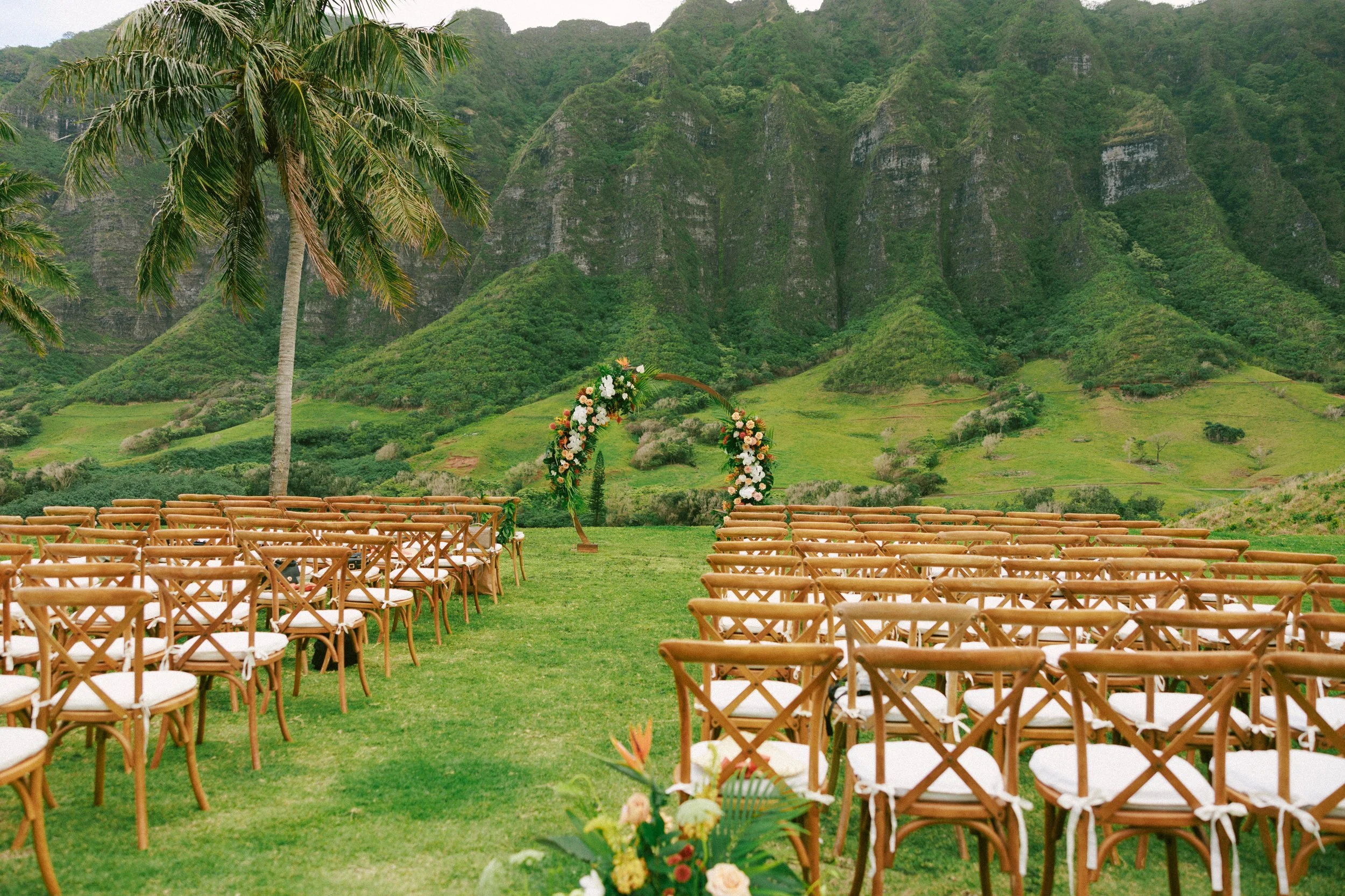 Outdoor wedding ceremony setup with white chairs arranged on lush green grass, surrounded by tropical palm trees and greenery under a partly cloudy sky.