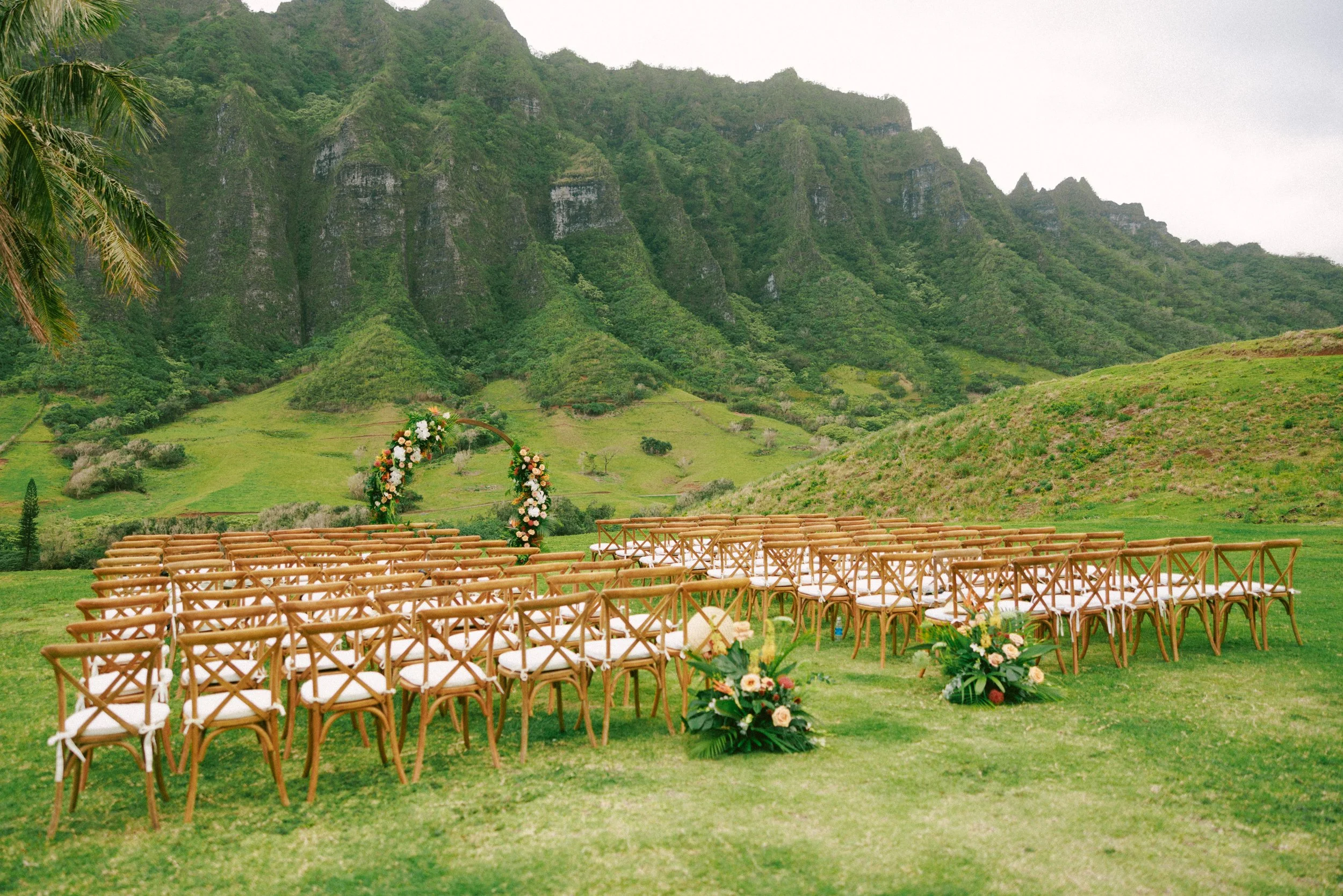 Outdoor wedding setup with rows of wooden chairs facing a floral arch in a scenic green valley surrounded by tall, lush mountains.