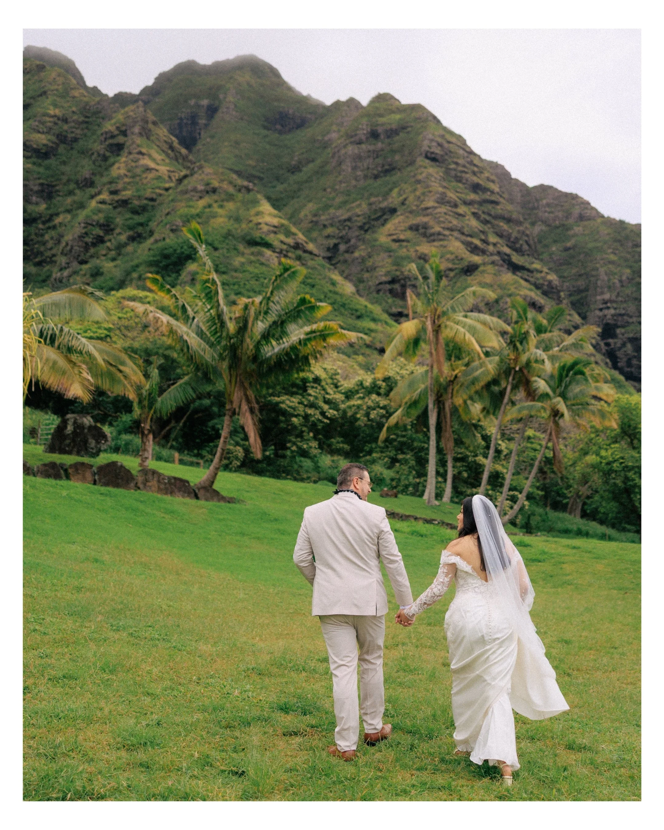 Bride and groom walk hand in hand across green lawn at Kualoa Ranch with Koʻolau mountains behind them during Oahu elopement wedding ceremony at Paliku Gardens
