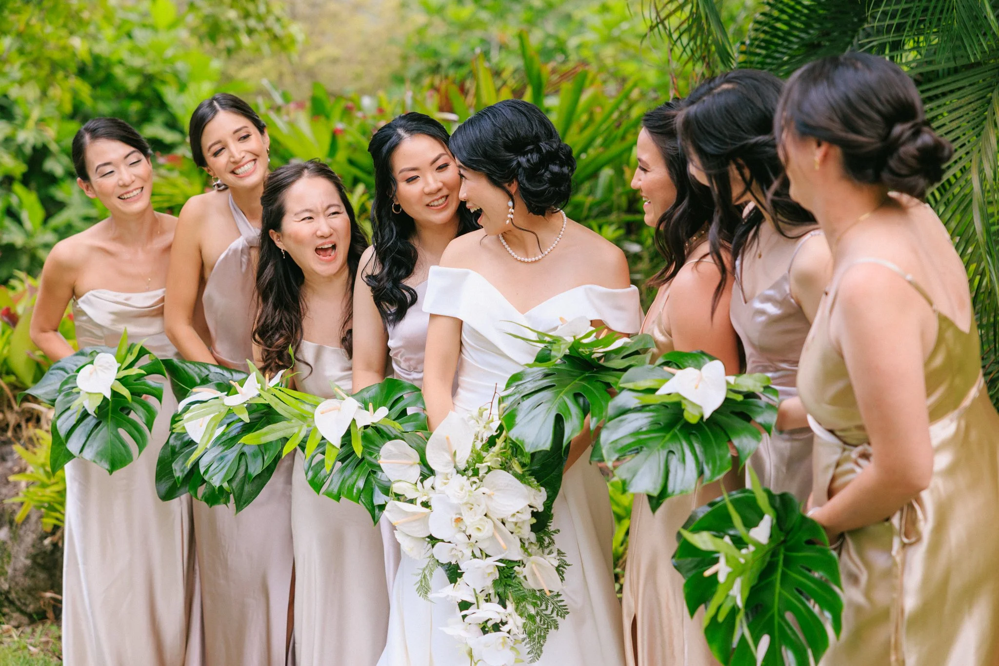 A bride in a white wedding dress surrounded by seven bridesmaids in beige dresses, all smiling and sharing a joyful moment outdoors with lush green tropical plants and large leaves in the background.