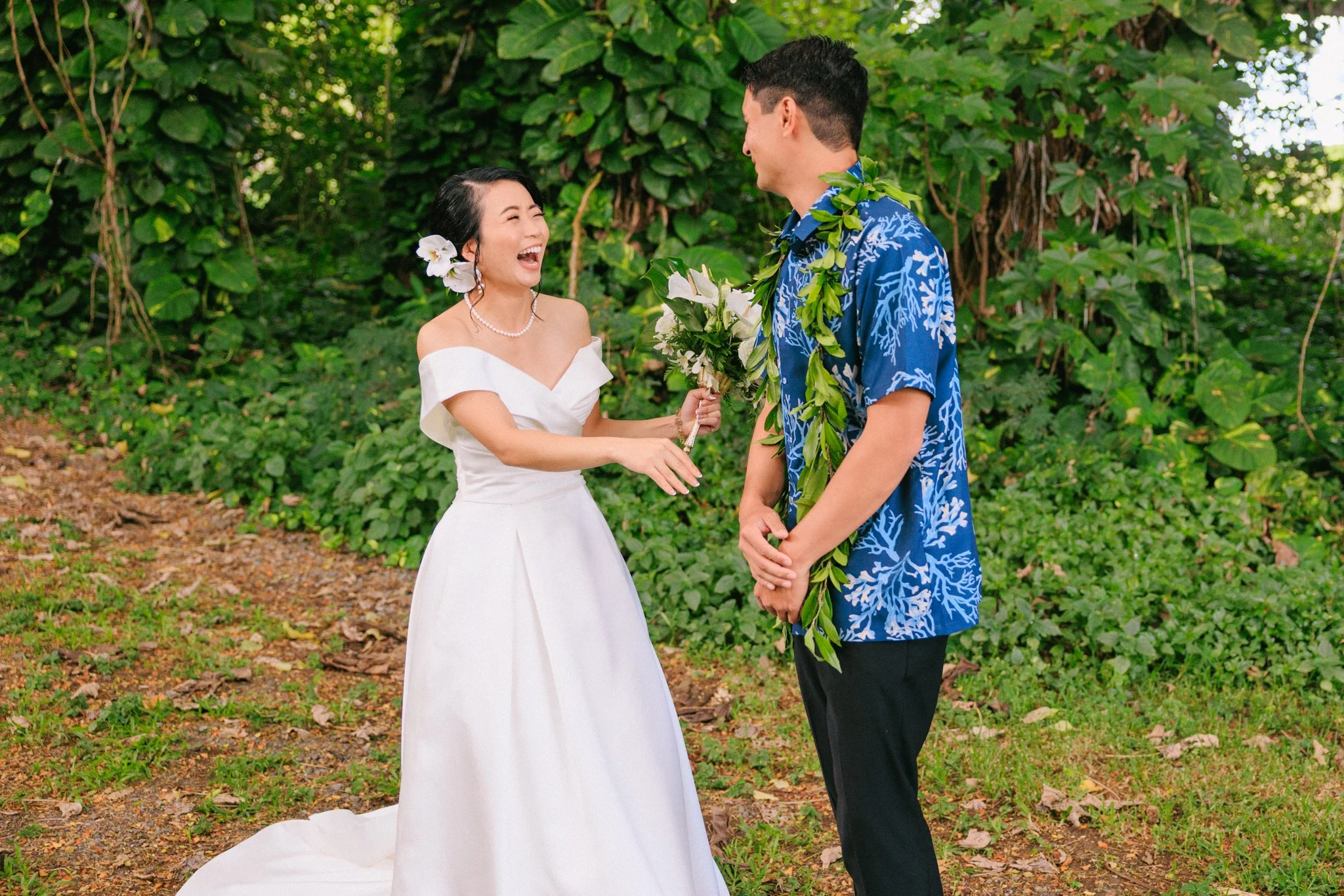 A woman in a white wedding dress laughing and holding a bouquet of white flowers, standing outdoors with a man in a blue Hawaiian shirt and black pants, who is wearing a green floral lei, in a lush green setting.