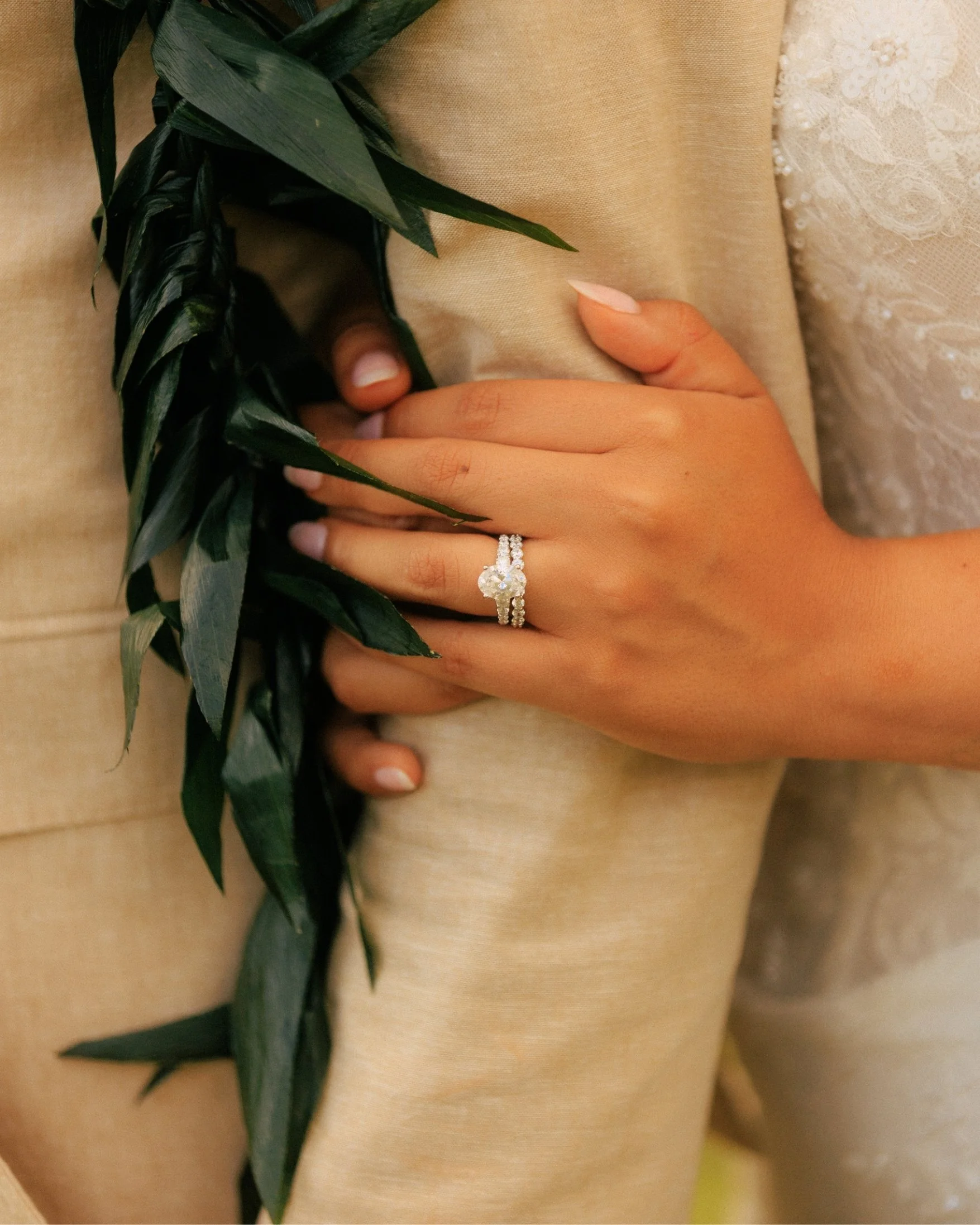 A close-up of Nicole's hand with her wedding ring resting on Ethan's tan suit beside his green leaf lei, a quiet detail from their elopement at Paliku Gardens, Kualoa Ranch, on Oahu, Hawaii.