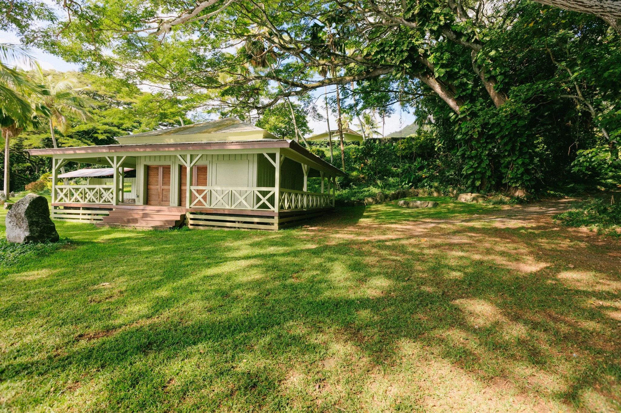 A small, green wooden house with stairs leading up to a porch, surrounded by lush green trees and grass, in a tropical setting.