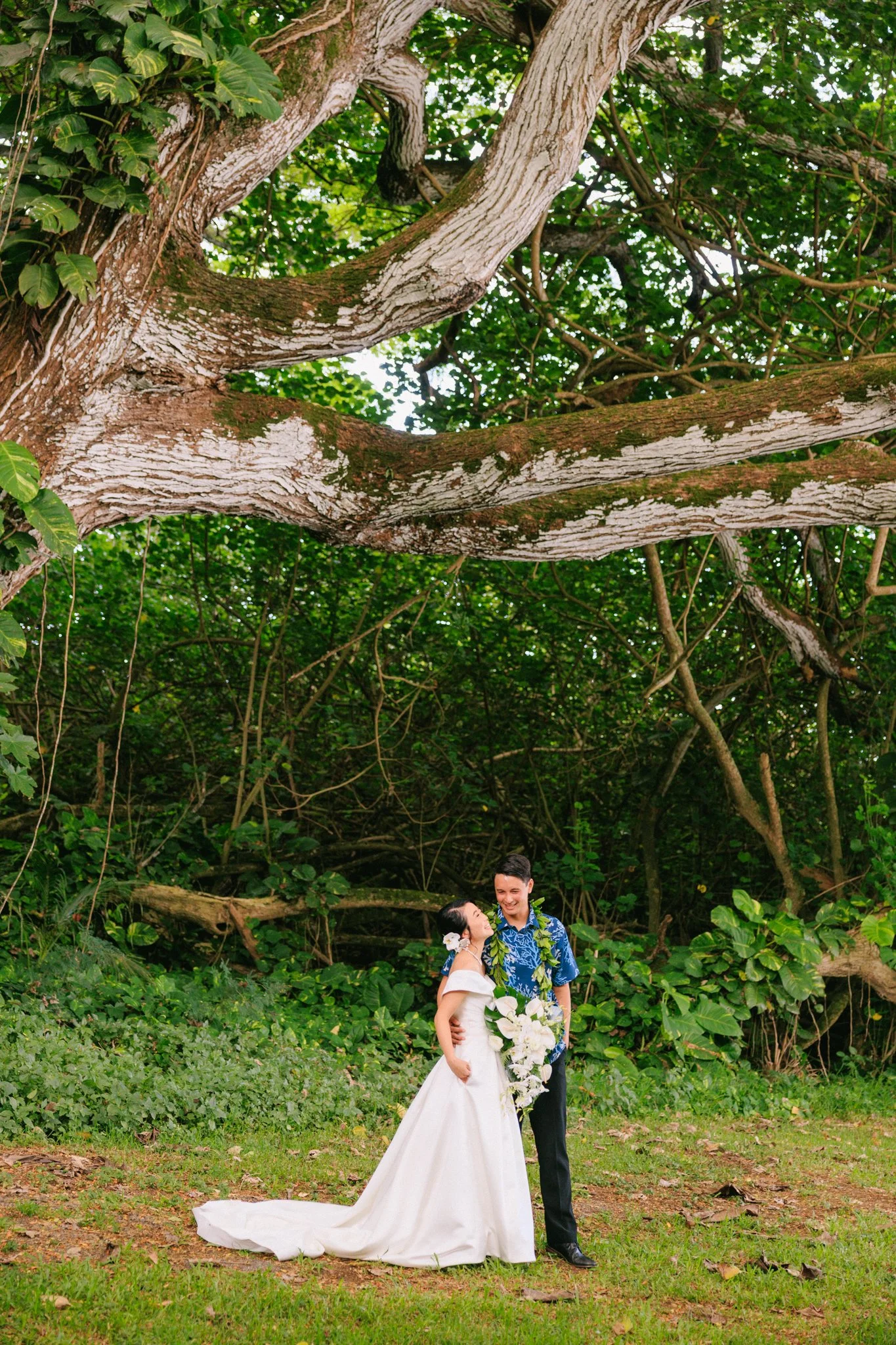 A couple in wedding attire standing on grass in front of lush green trees, sharing a joyful moment. The bride is wearing a white wedding gown and holding a bouquet of white flowers, while the groom is dressed in a blue shirt with a floral pattern. Th