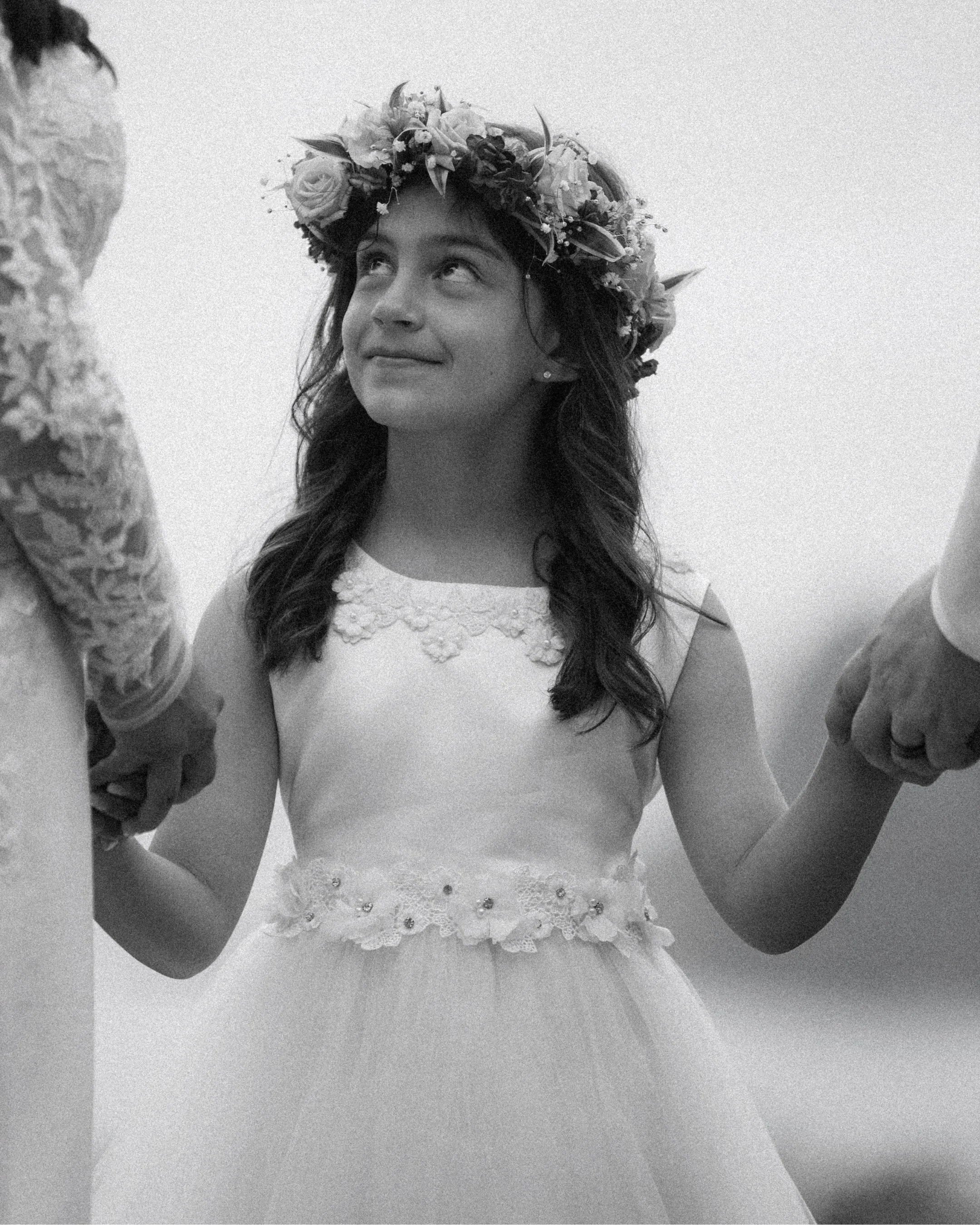 Flower girl in white dress and flower crown looks up while holding hands during emotional family wedding ceremony at Kualoa Ranch Oahu.