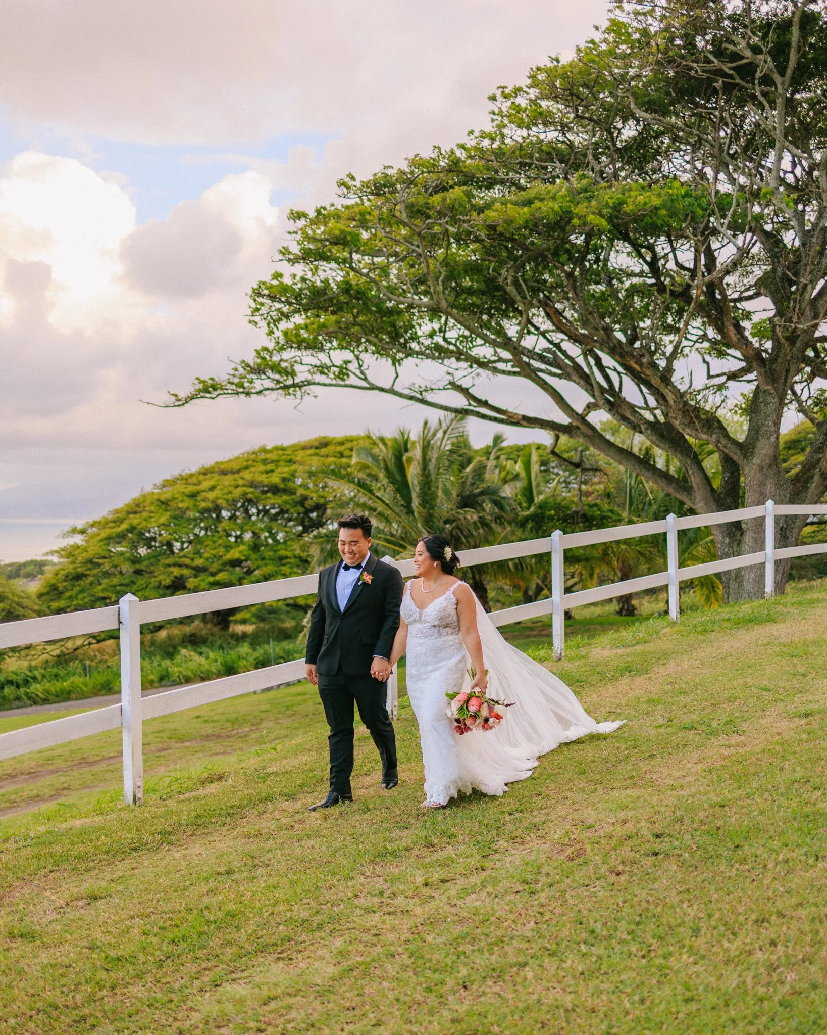 A newlywed couple walking hand-in-hand outdoors on grass, smiling. The bride is holding a bouquet of flowers and wearing a white wedding gown; the groom is in a black suit with a bow tie. Large trees and a white fence are in the background under a pa