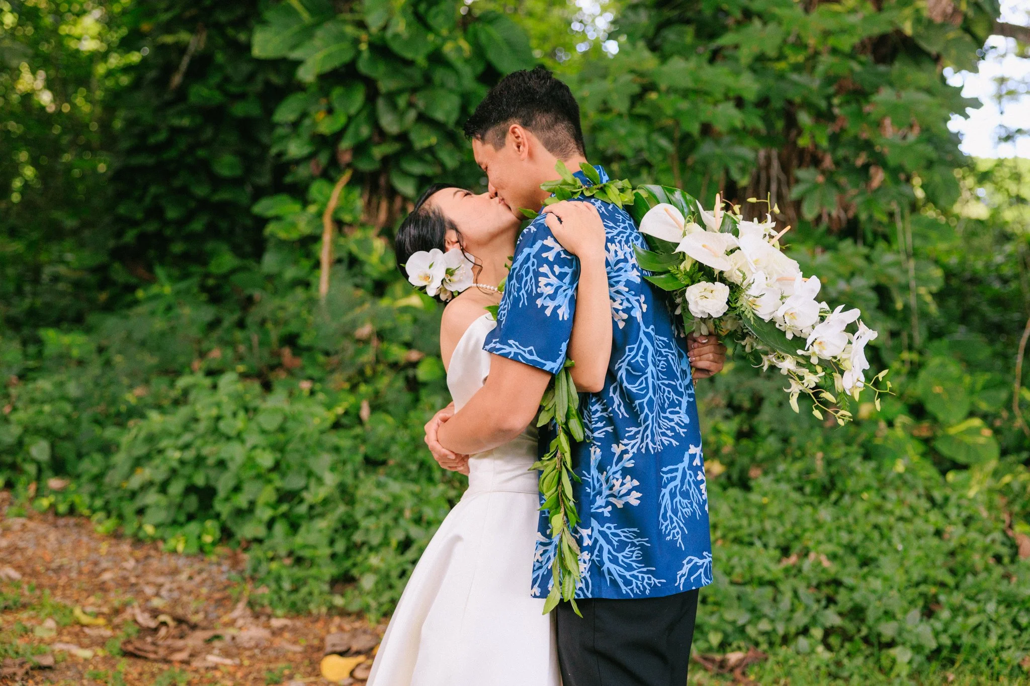 A couple kissing outdoors, with the woman holding a large bouquet of white flowers and the man wearing a blue patterned shirt, surrounded by lush green foliage.