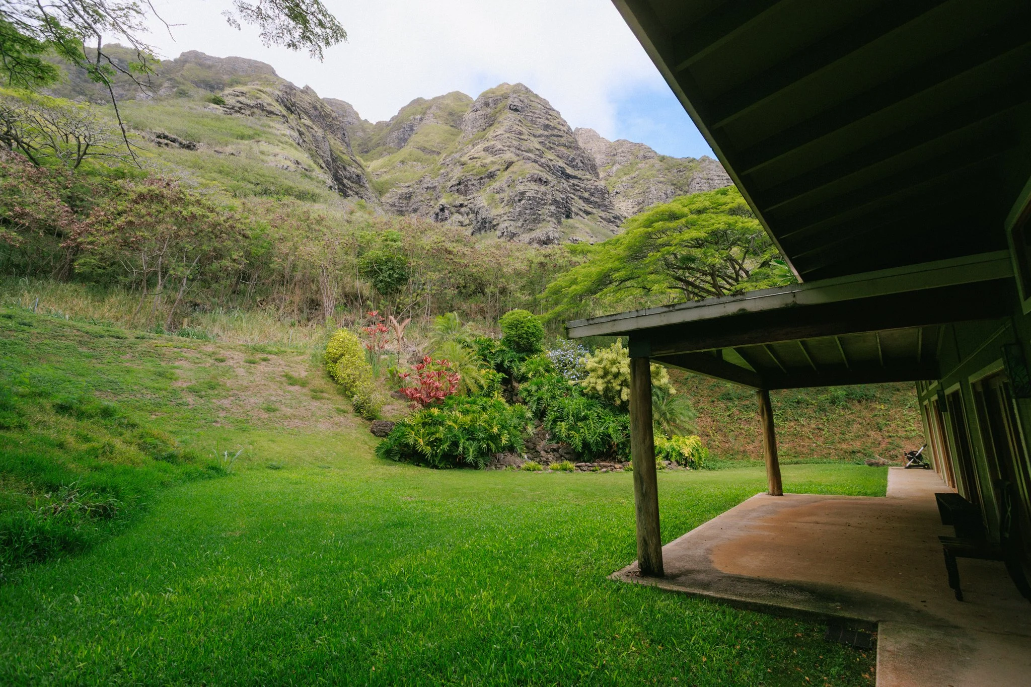 Lush green lawn with hillside and mountain backdrop, covered patio with support posts, steps leading to a walkway, and tropical plants along the hillside