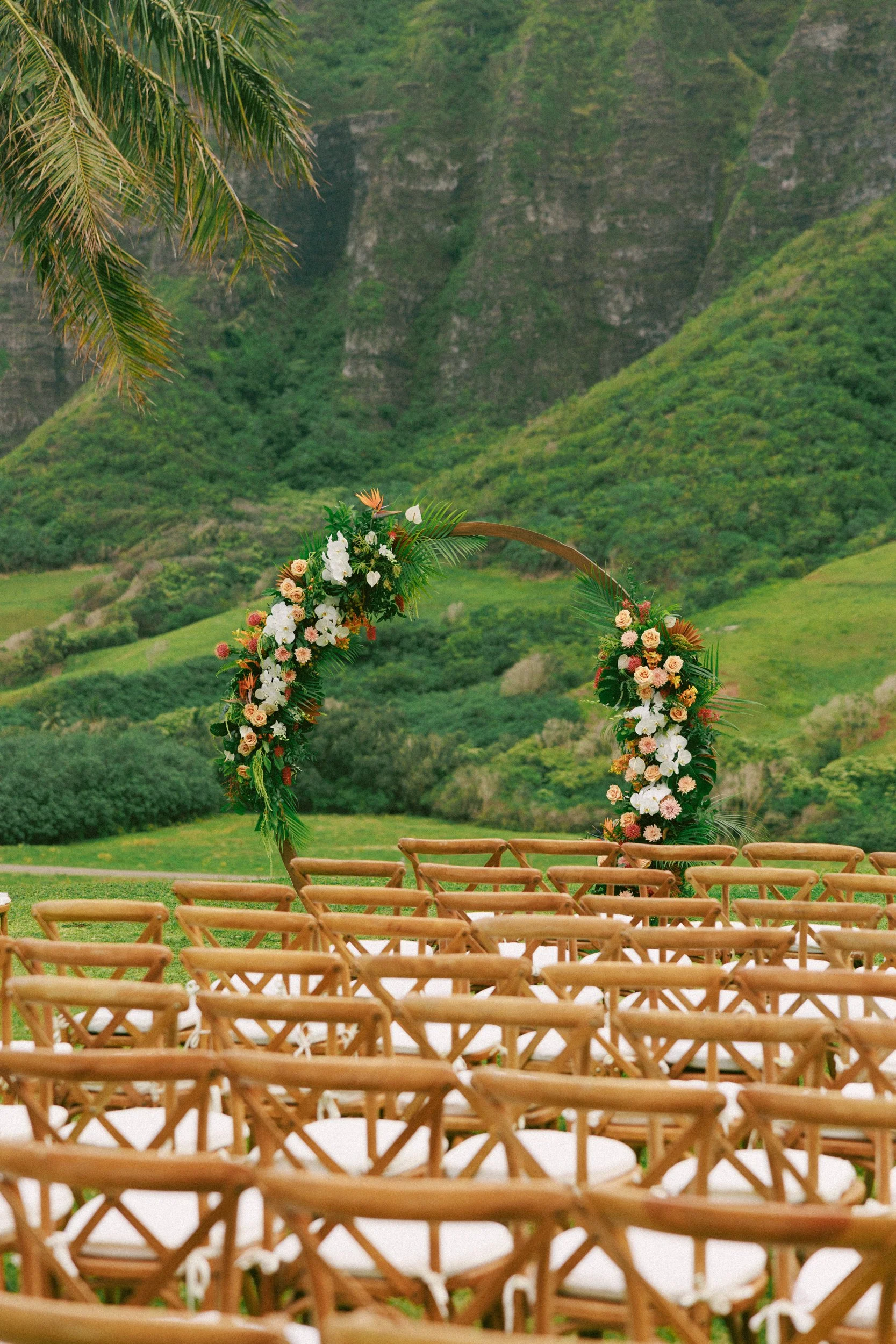 A wedding arch decorated with flowers and greenery in an outdoor setting with lush green hills in the background, and rows of wooden chairs with white cushions in the foreground.