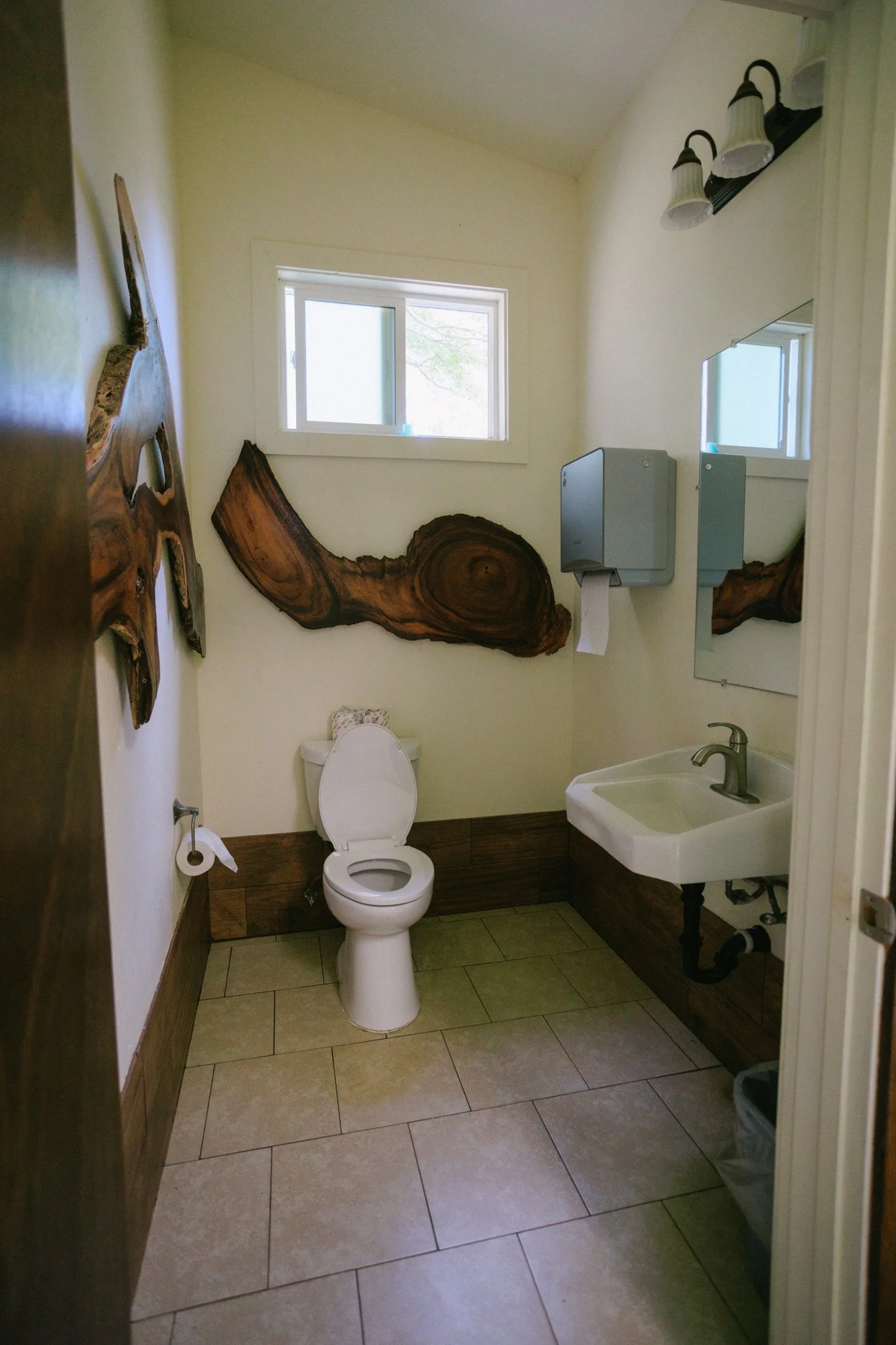 Small bathroom with a toilet, a small sink, wooden accents, and decorative wooden pieces on the wall. A paper towel dispenser and a mirror are above the sink, and there is a small window letting in natural light.