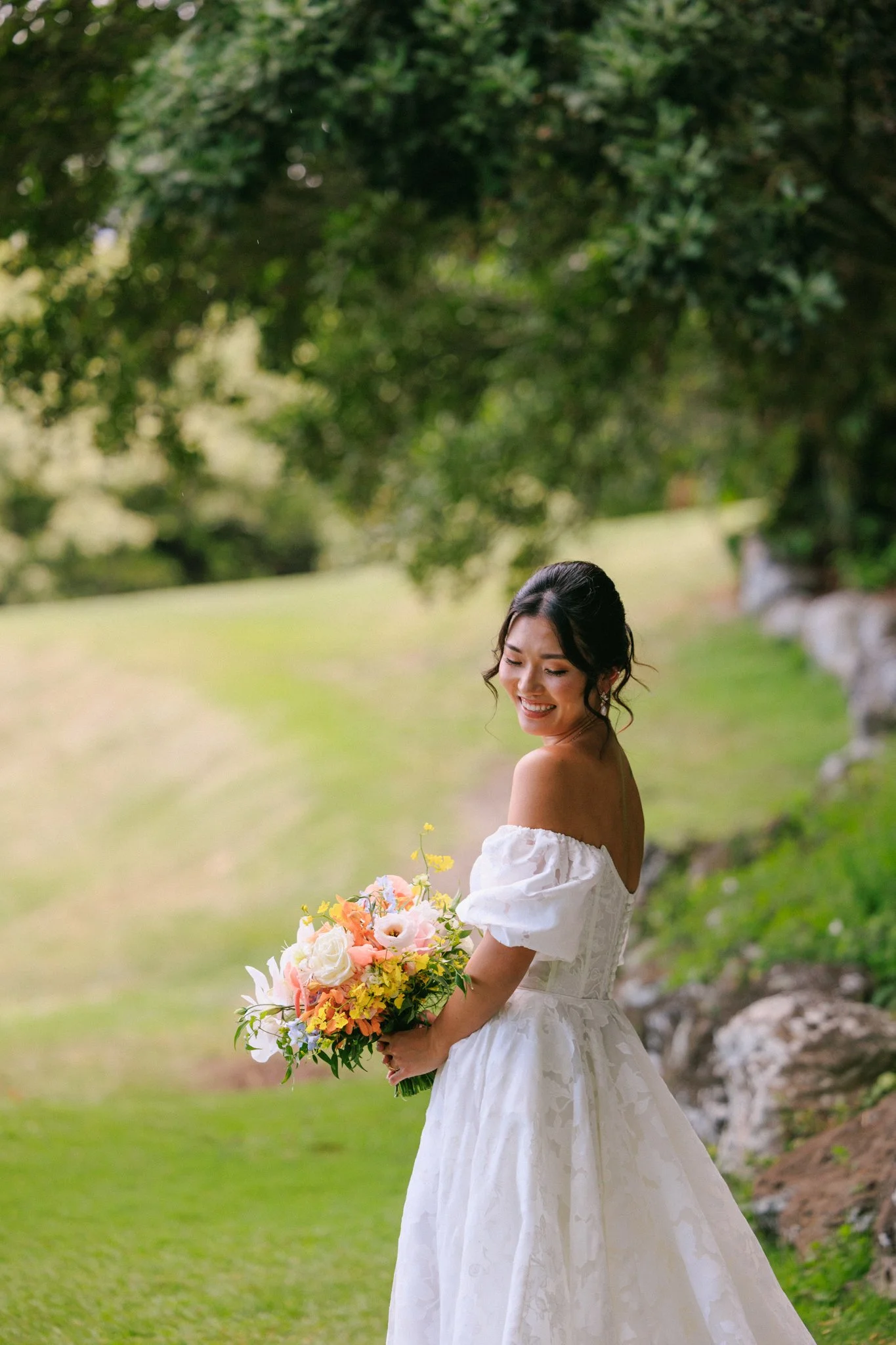 A woman in a white off-shoulder dress holding a colorful bouquet of flowers, smiling outdoors with trees and grass in the background.