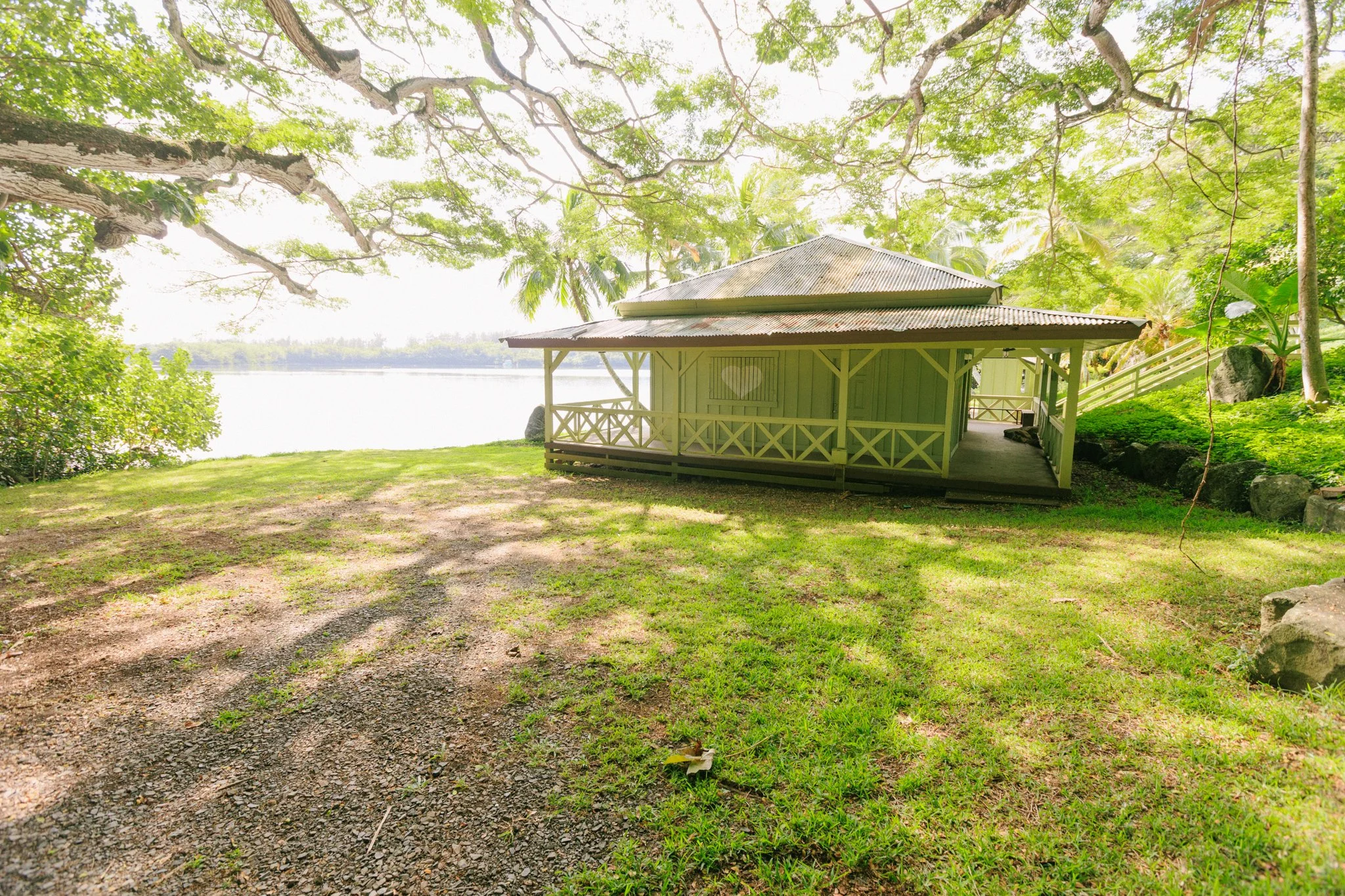 A small light green wooden cabin with a heart on its wall situated near a body of water, surrounded by lush green trees in a peaceful, shaded tropical setting.