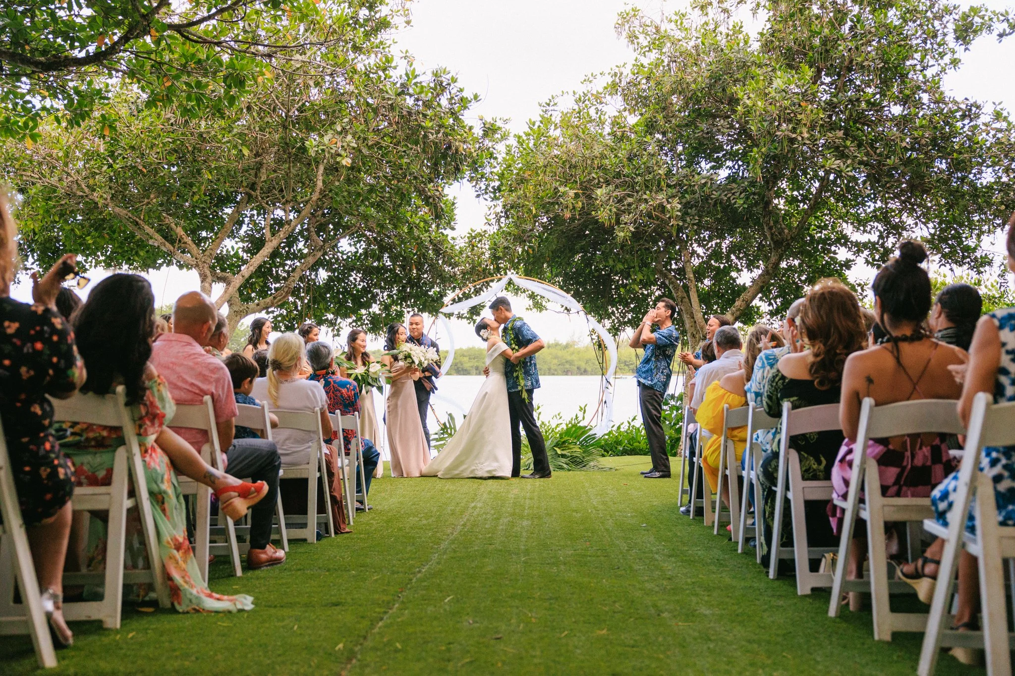 A wedding ceremony taking place outdoors between a bride and groom under a circular arch, with guests seated on either side and lush greenery and trees in the background.