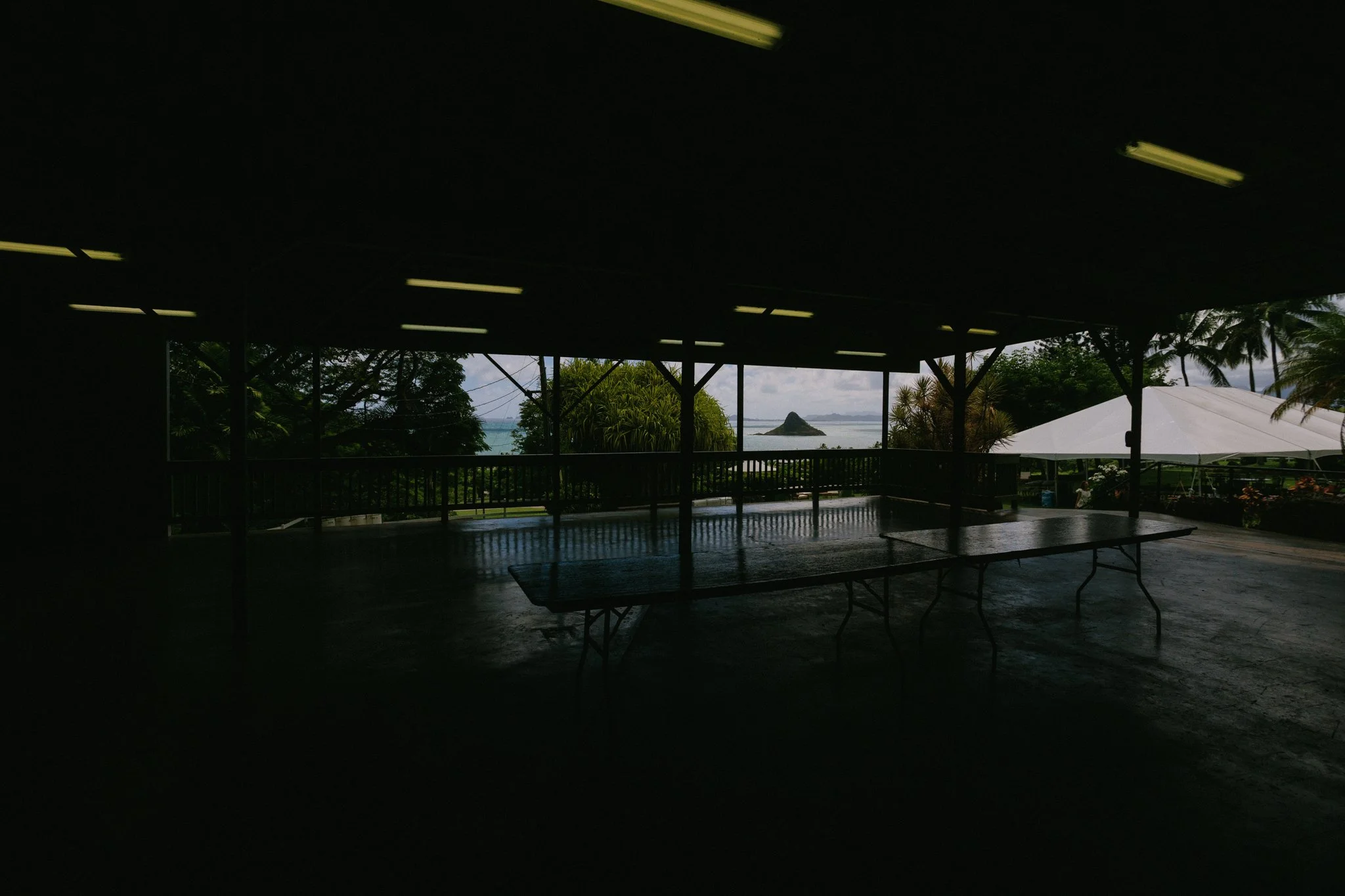 Dark interior space with tables and bright outdoor view of trees, water, and an island through large open windows.