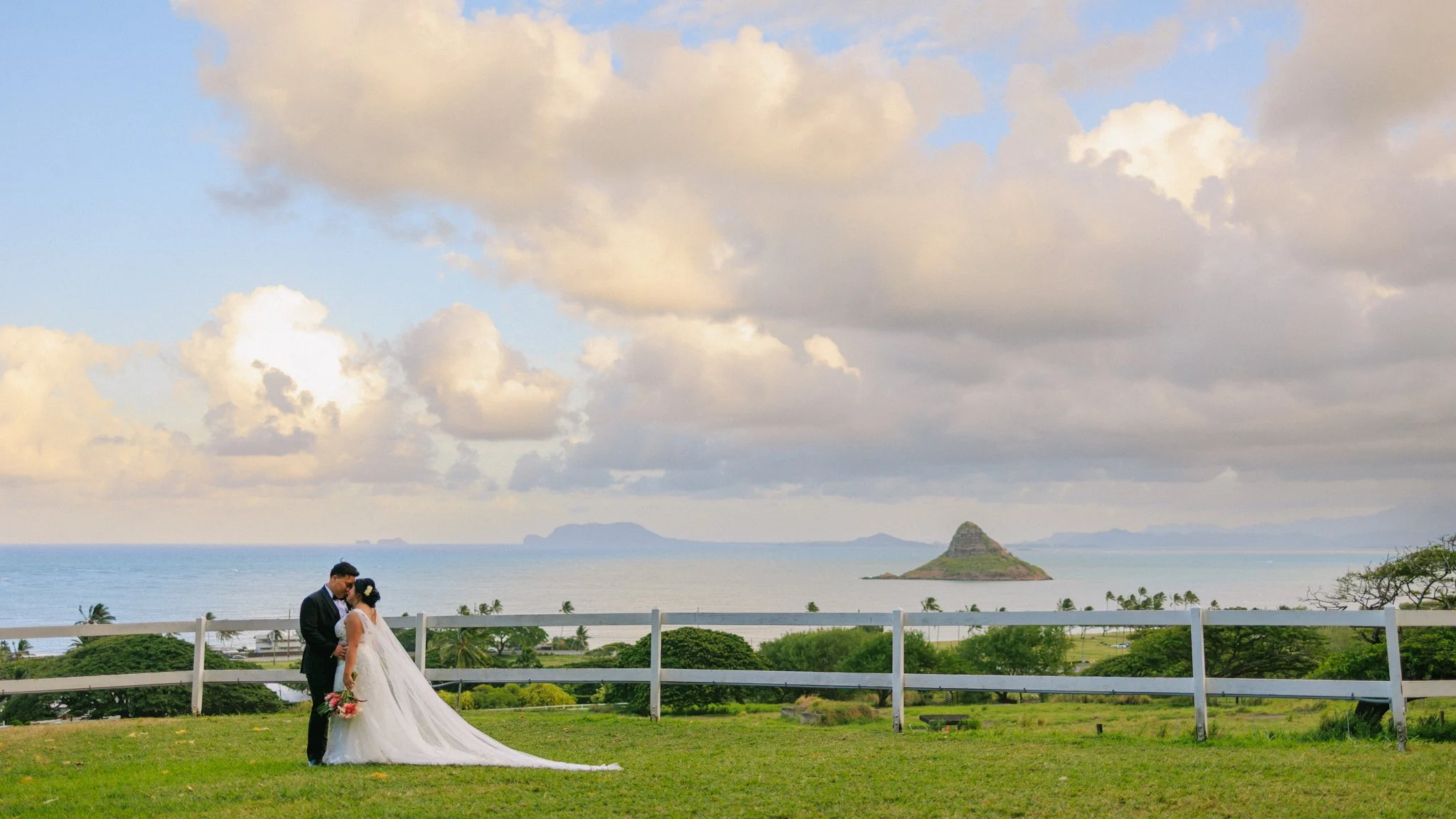 A bride in a white wedding dress and a groom in a black suit kiss outdoors on a grassy area with a white fence, overlooking the ocean, islands, and a partly cloudy sky.