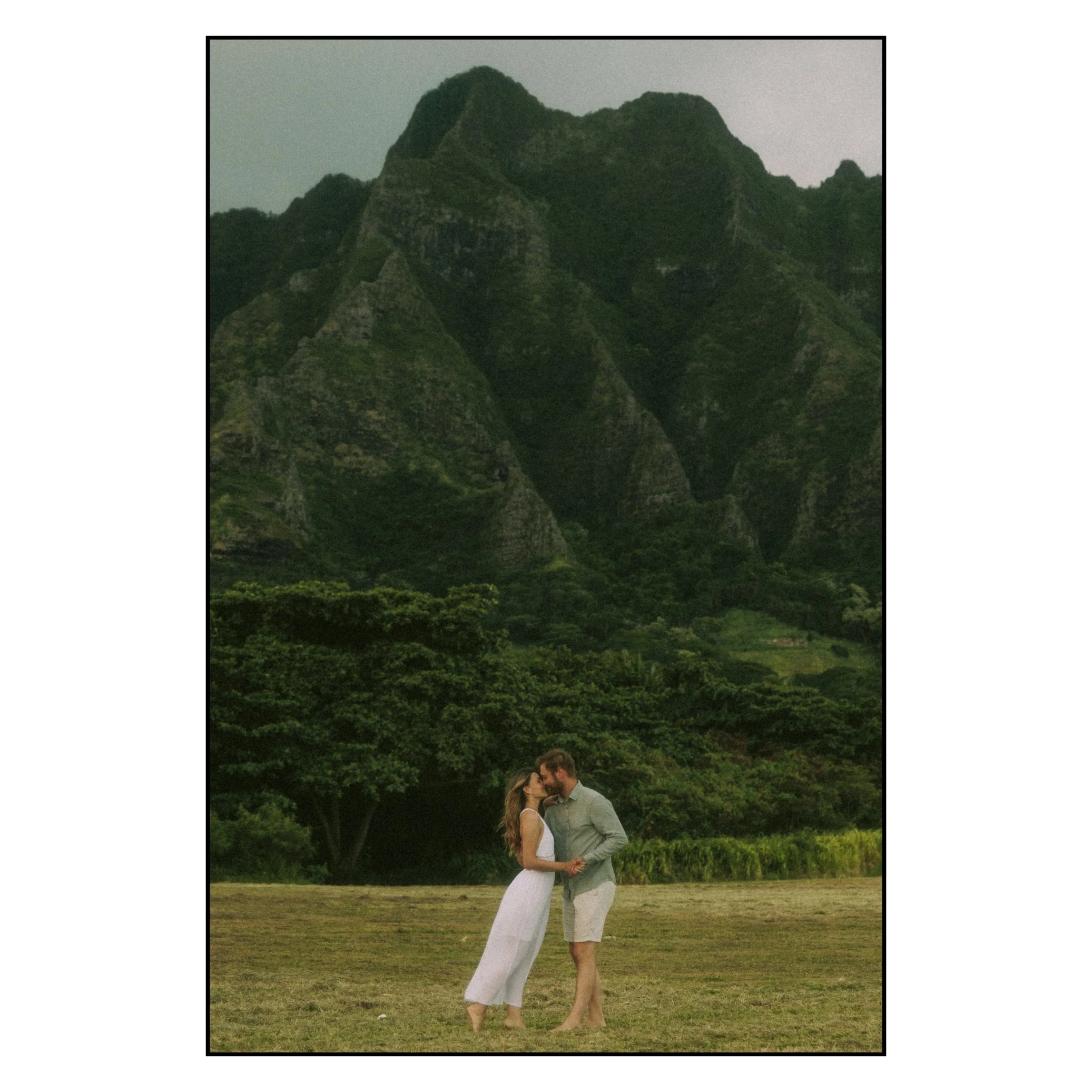 Couple sharing a quiet kiss in an open field beneath the towering green mountains at Kualoa Regional Park, Oahu.