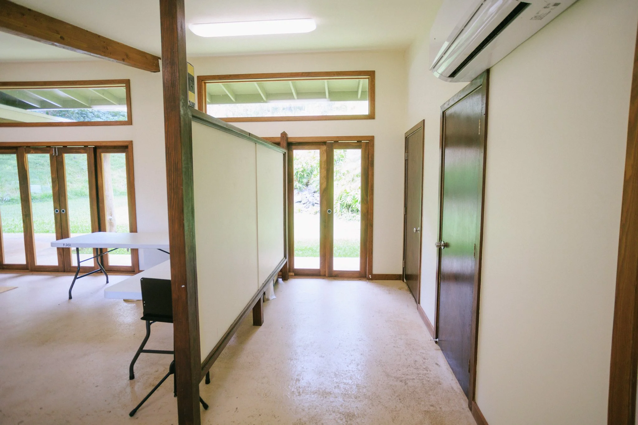 Interior view of a room with large windows, glass door, wooden frames, a split air conditioning unit, and folding tables with black chairs.
