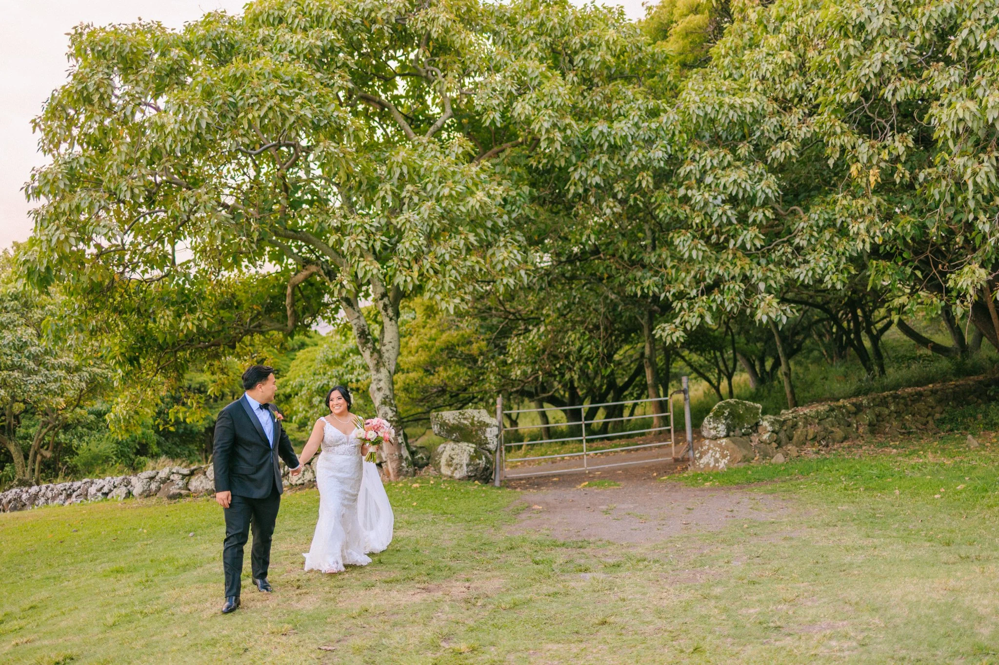 Bride and groom walking hand in hand outdoors in a park surrounded by green trees and grass, with a dirt path and a metal gate in the background.