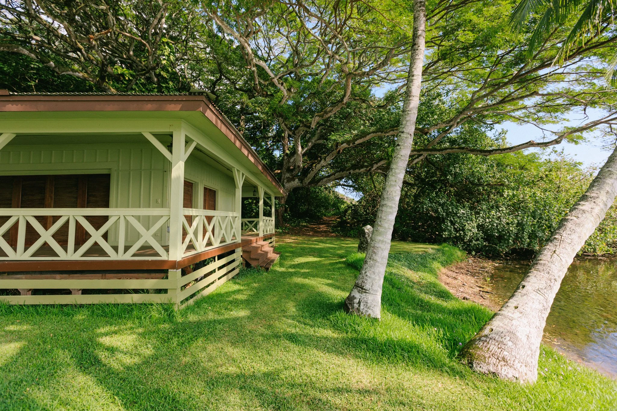 A house with a green exterior, brown trim, and a veranda with white cross railing, located along a grassy area with mature trees and waterfront water.