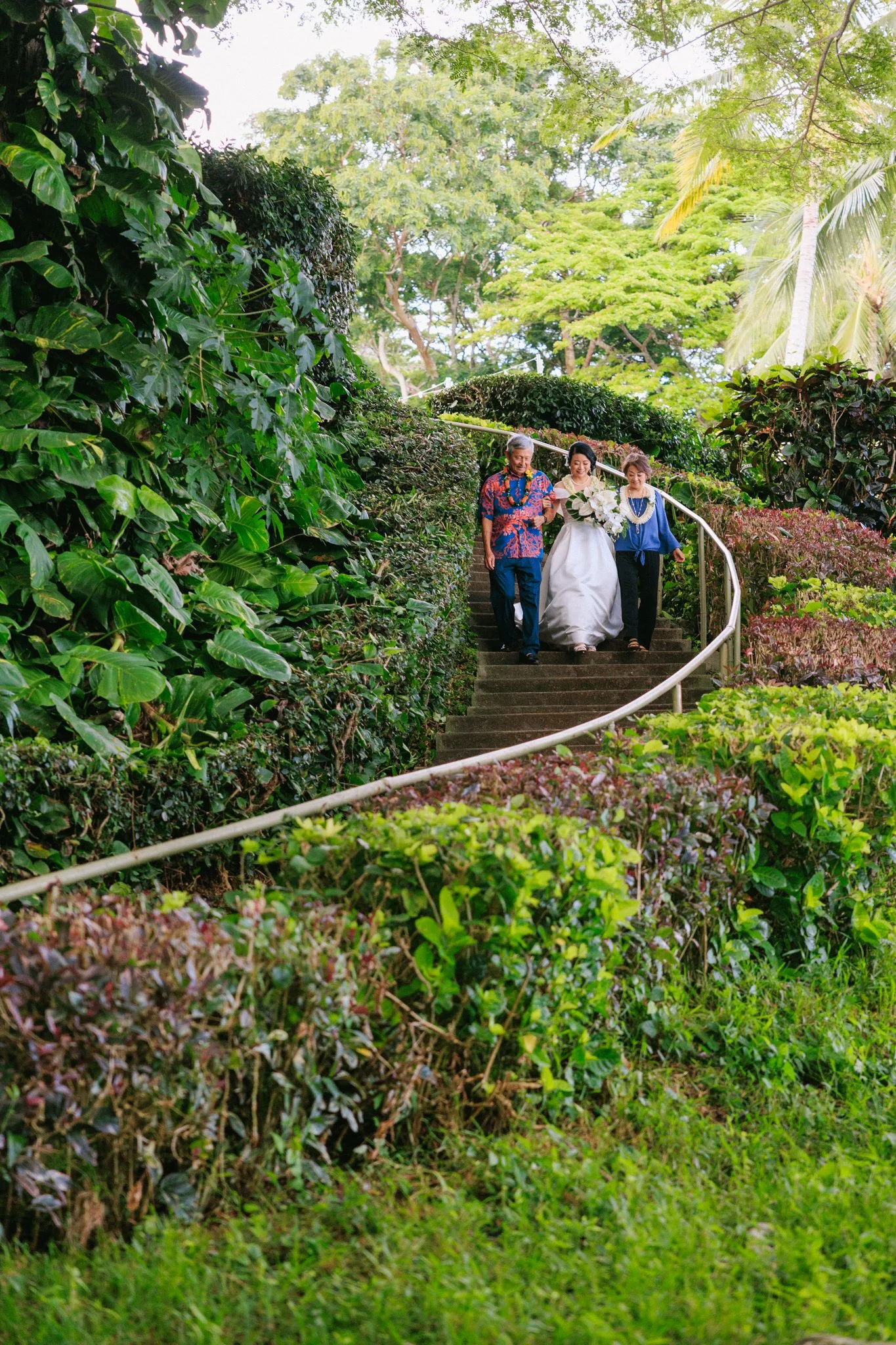 A bride in a white wedding dress walking down outdoor stairs with two women, one elderly, and a man, all dressed in colorful shirts, surrounded by lush green plants.