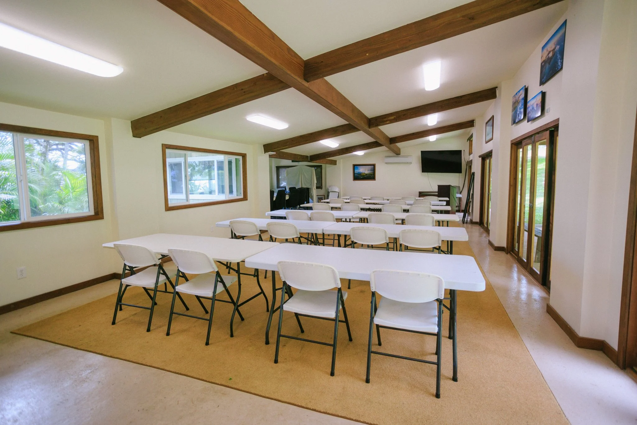 An empty conference room with multiple white tables and chairs, large windows, and a mounted TV on the wall.