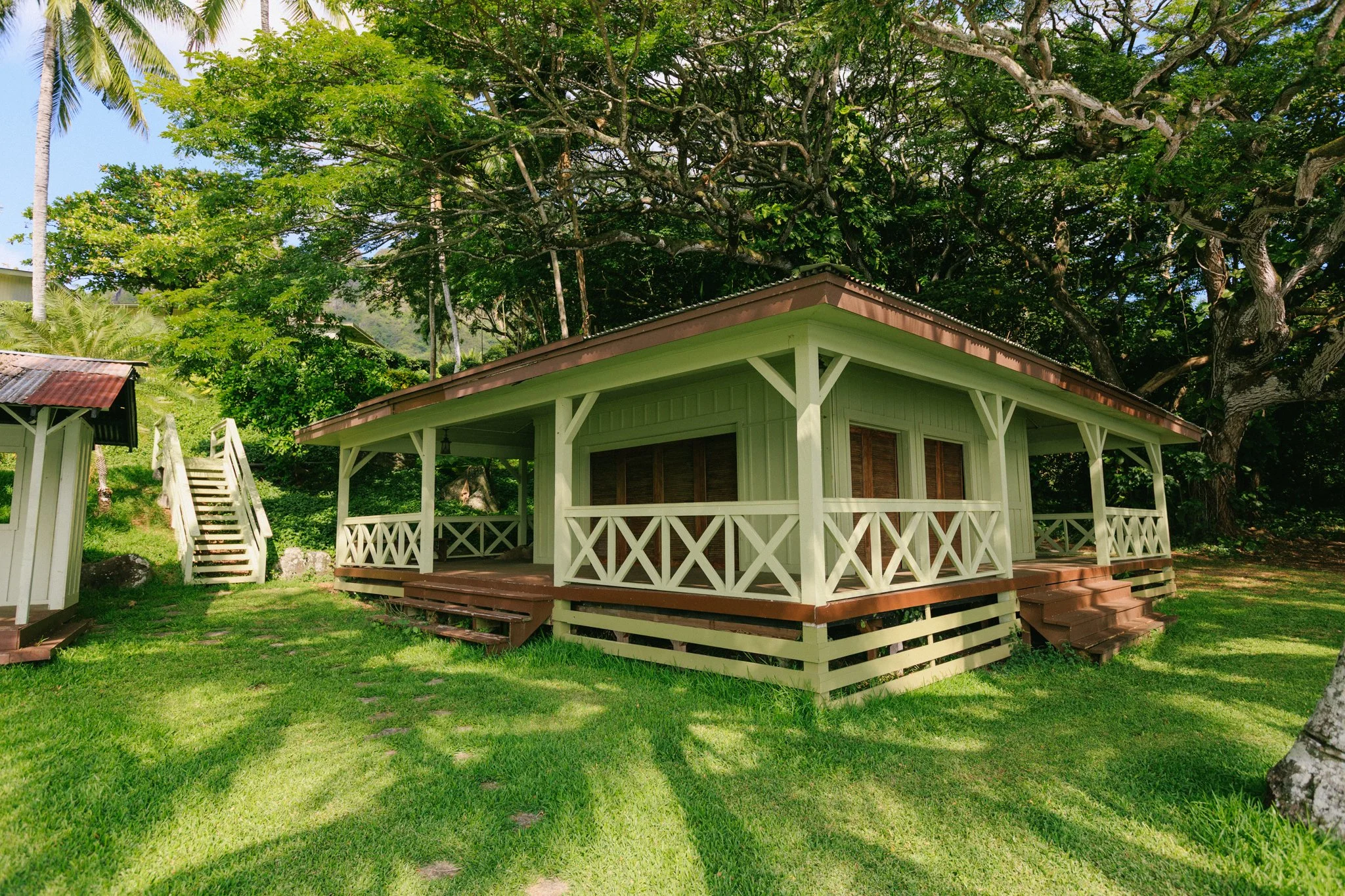A small, light green house with a porch surrounded by lush green trees and grass.