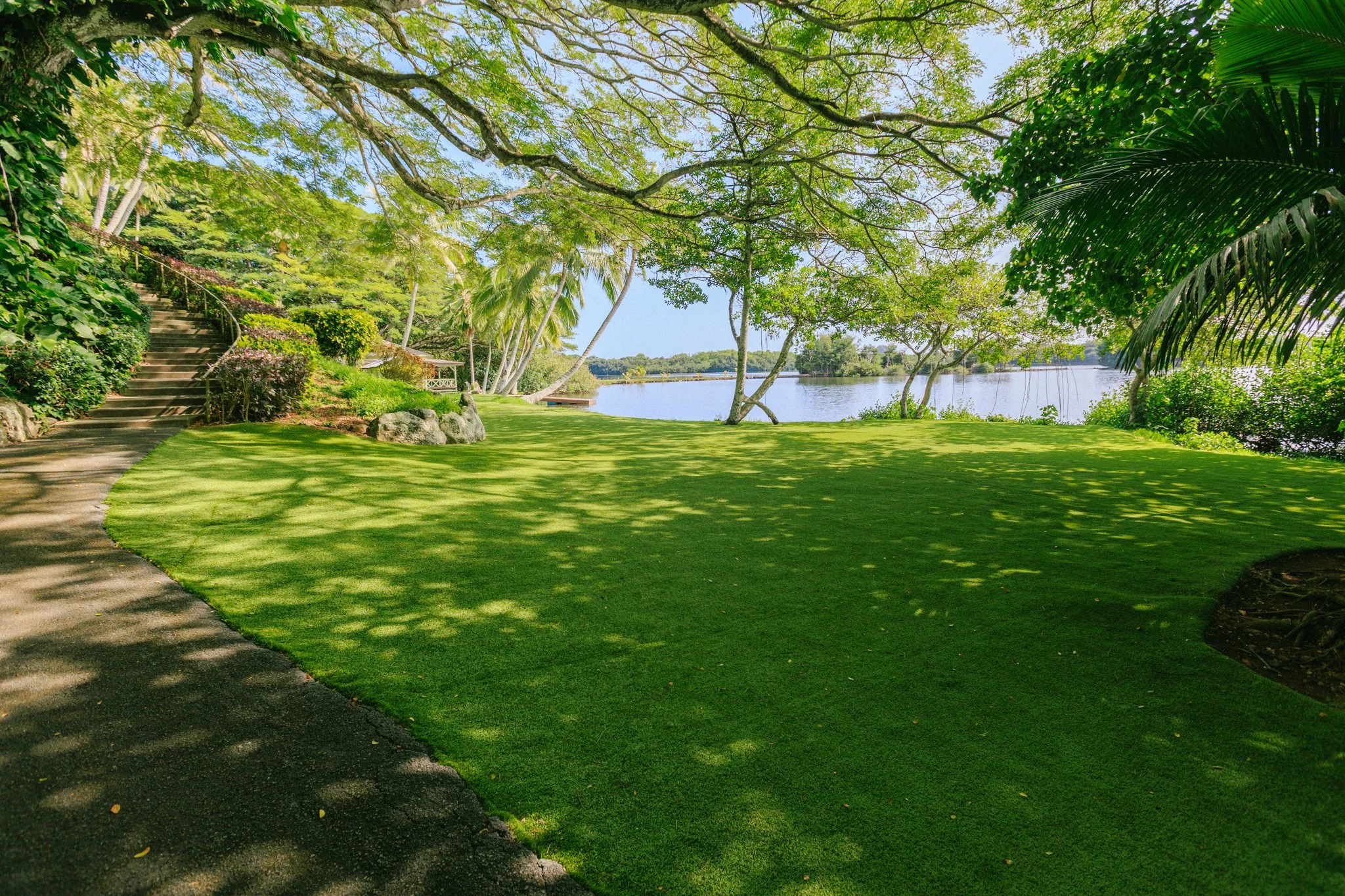Lush green backyard with a curved stone pathway, overlooking a body of water, shaded by large trees.