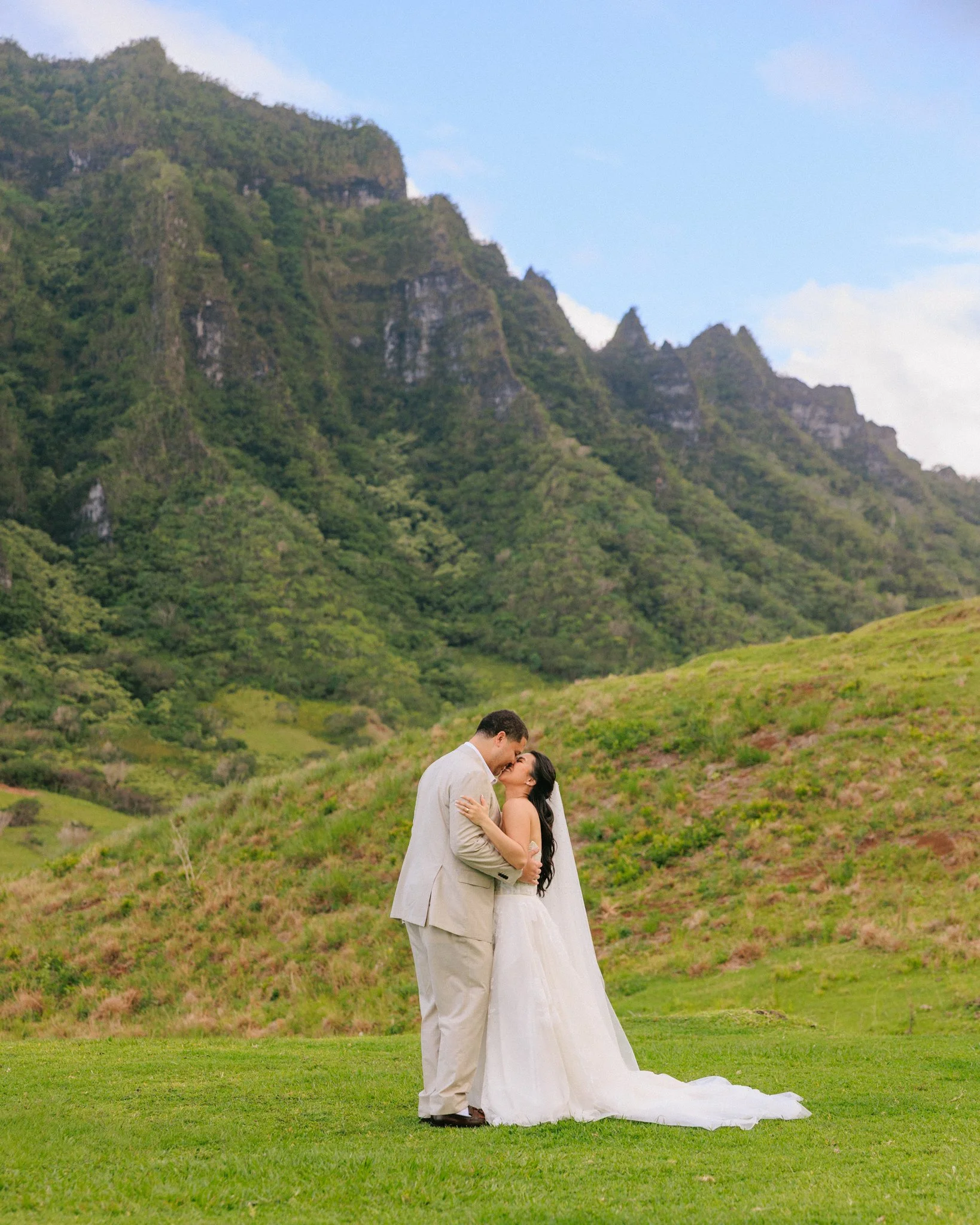 A bride and groom kissing outdoors on green grass with green mountains and blue sky in the background.