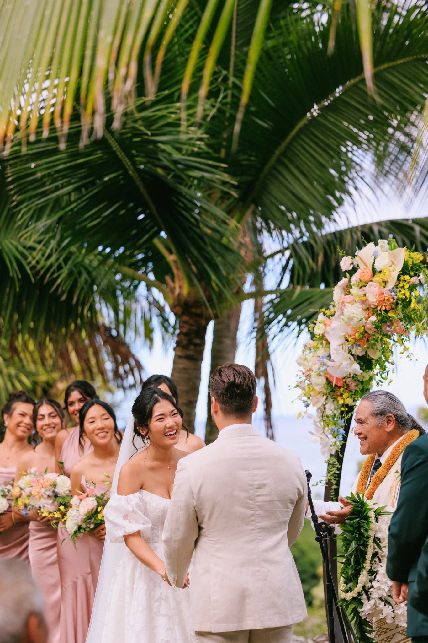 A wedding ceremony taking place outdoors under palm trees, with the bride and groom exchanging vows, surrounded by bridesmaids and an officiant, with floral decorations.