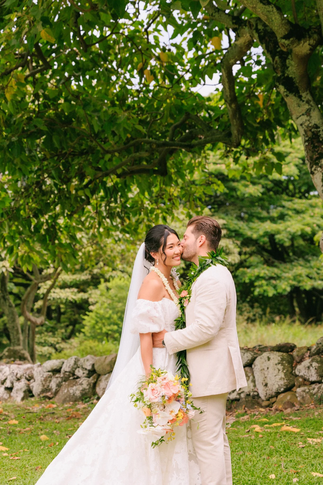 A bride and groom embrace outdoors, with the groom kissing the bride's cheek, surrounded by lush green trees and grass.