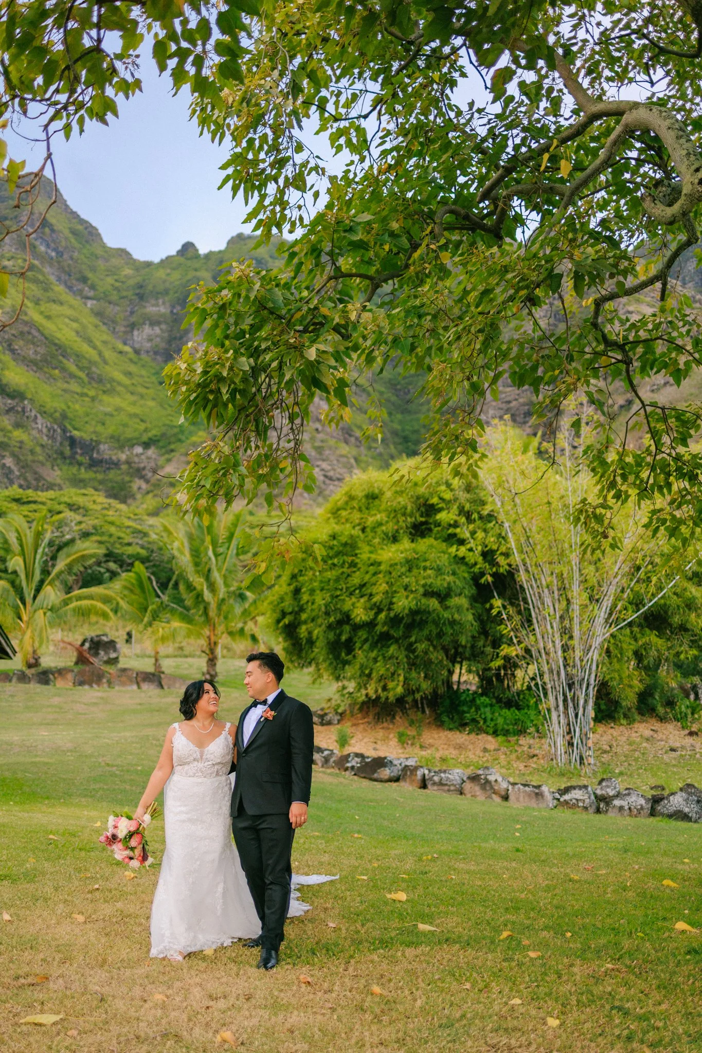 A newlywed couple walking hand in hand on a grassy area with large trees and mountains in the background, bride holding a bouquet of flowers.