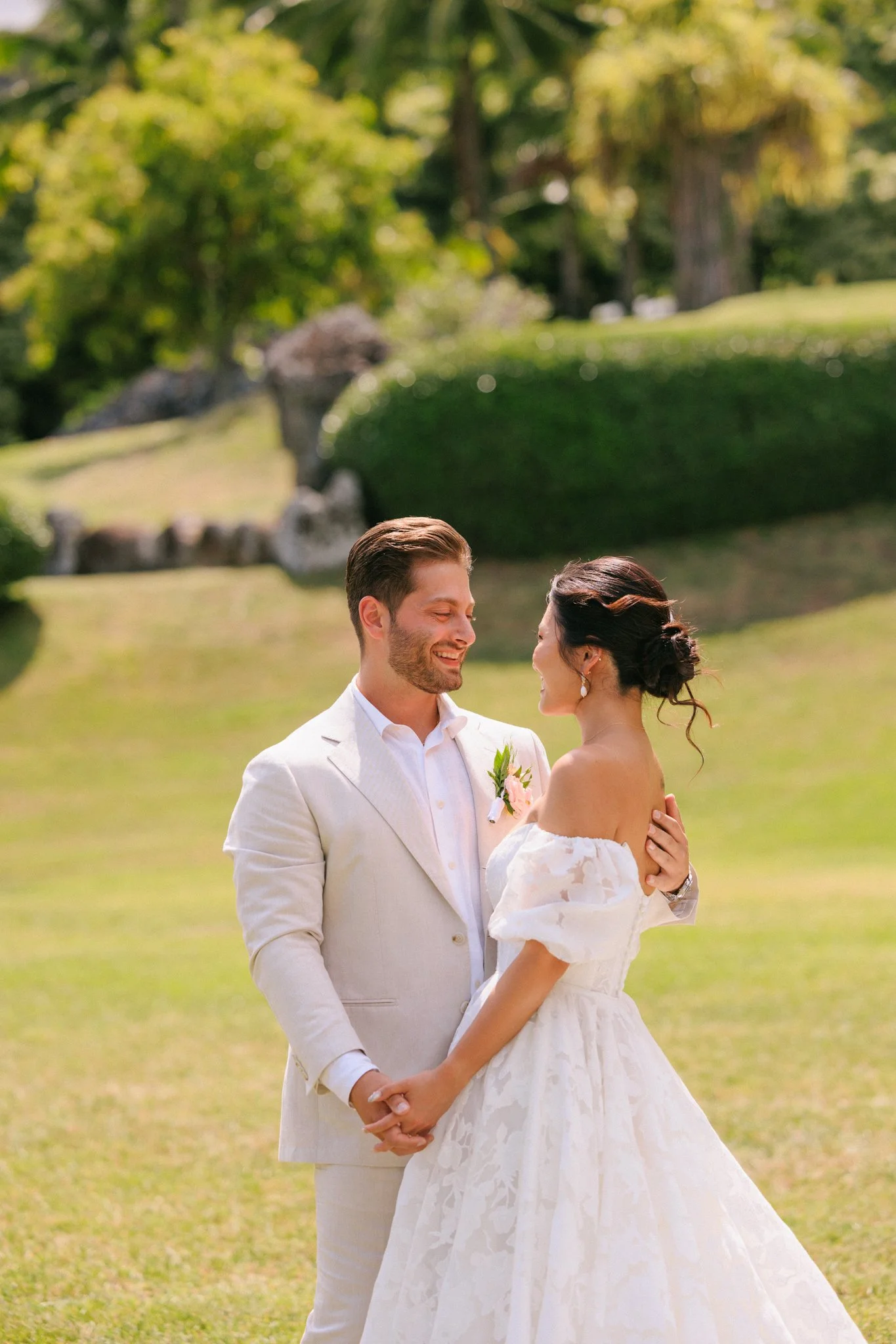 A bride and groom holding hands and gazing at each other outdoors in a grassy park, with trees and rocks in the background, on a sunny day.