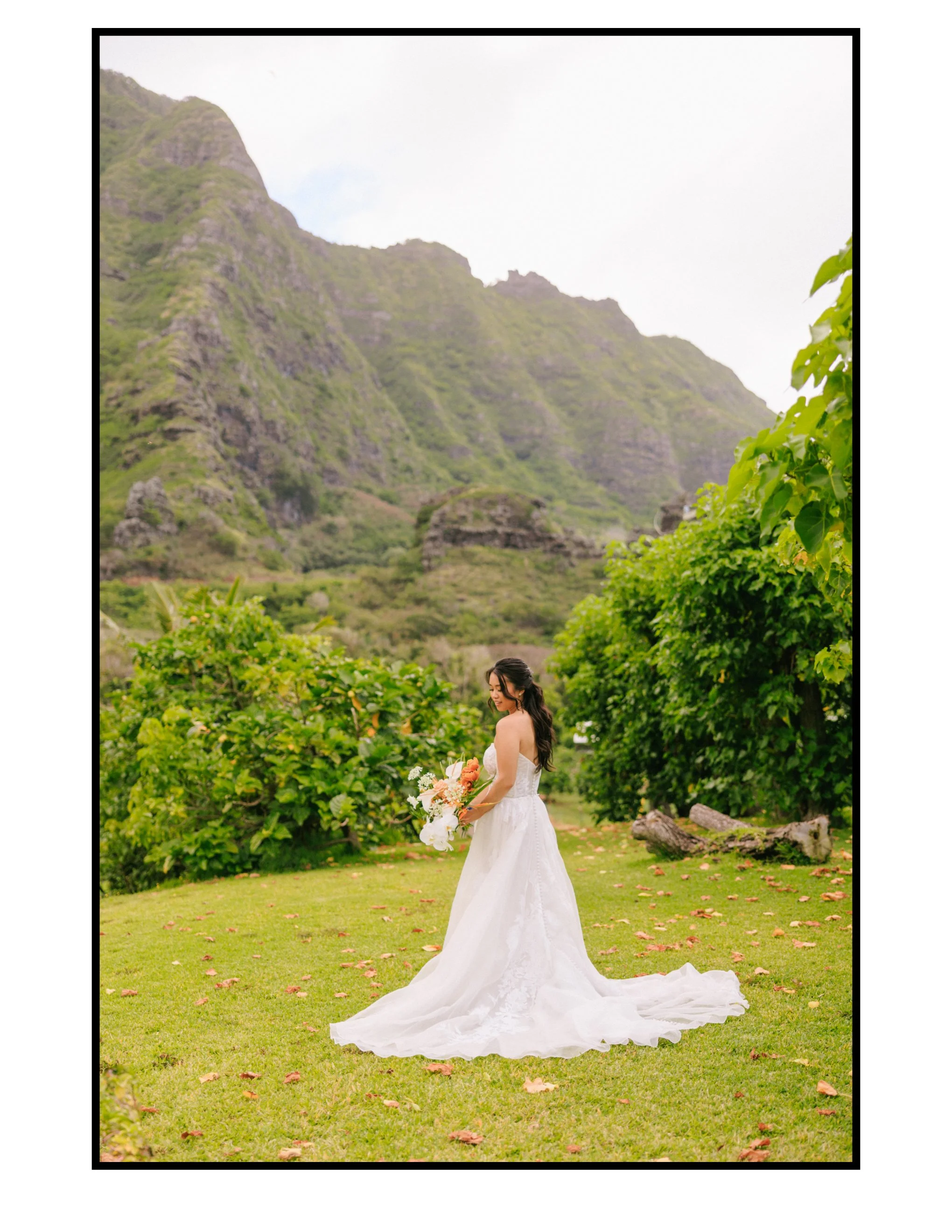 Bride in a white wedding dress holding a bouquet, standing on a grassy field surrounded by green trees with a mountainous landscape in the background.