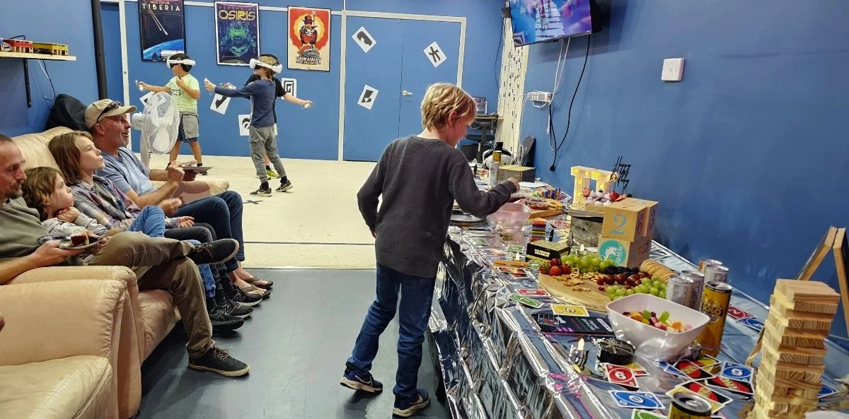 birthday party at DropBear Escape Rooms Pakenham.  boy choosing food from a table, other kids in the background playing VR. adults sitting on the couch watching othe kids on the tv.