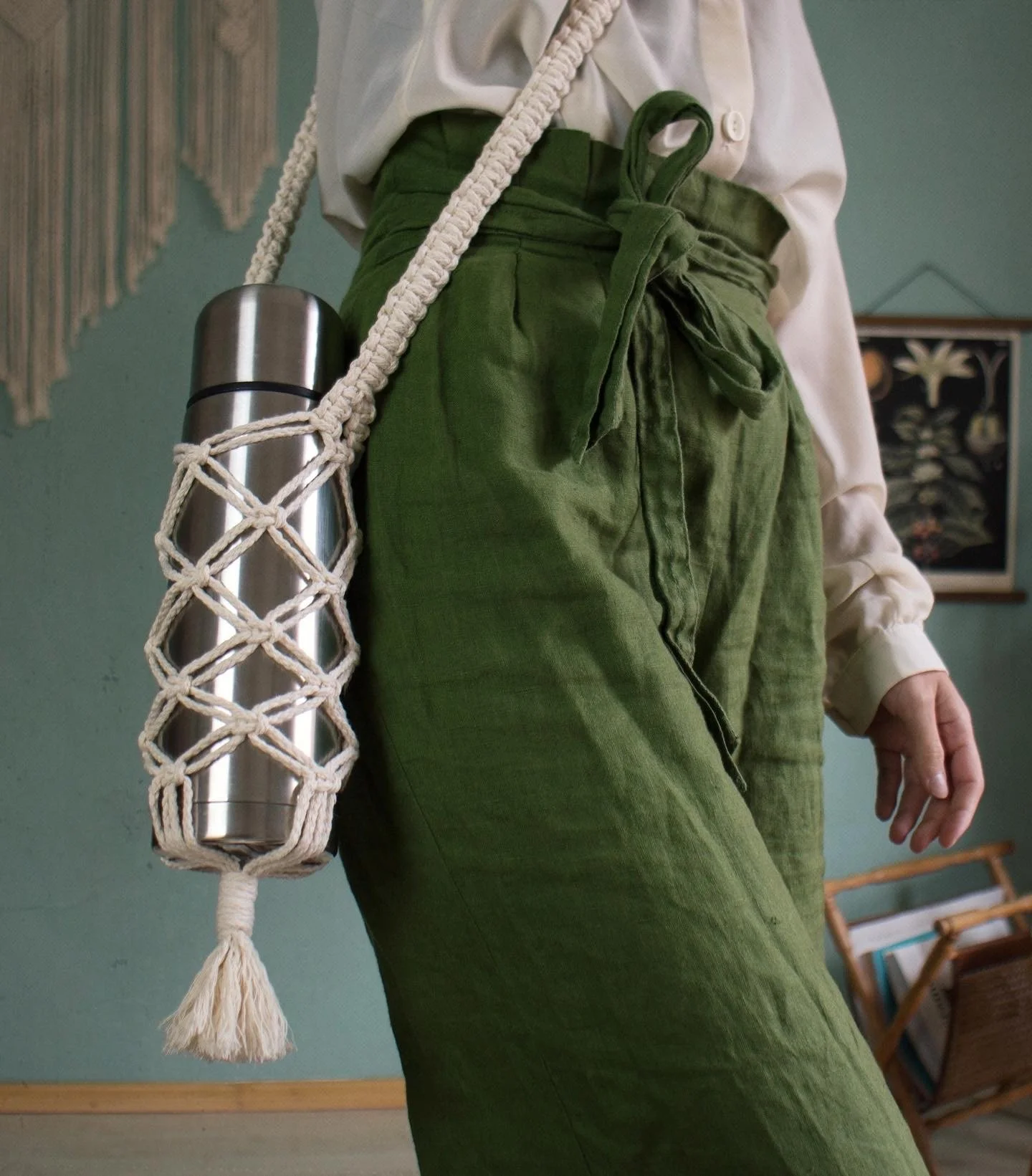 Du siehst auf dem Bild einen Ausschnitt einer Frau, die eine grüne Hose trägt. An ihrer Schulter hängt ein aus Makrameegarn geknüpfter Flaschenhalter mit einer silbernen Flasche.